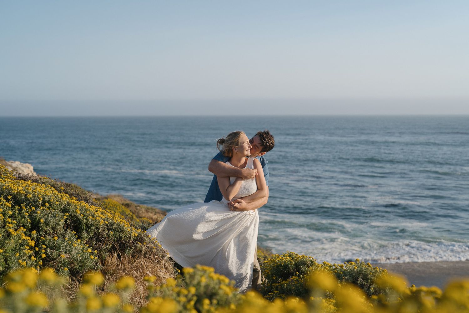 A couple shares a romantic moment on a coastal cliff adorned with yellow wildflowers overlooking the Pacific Ocean.