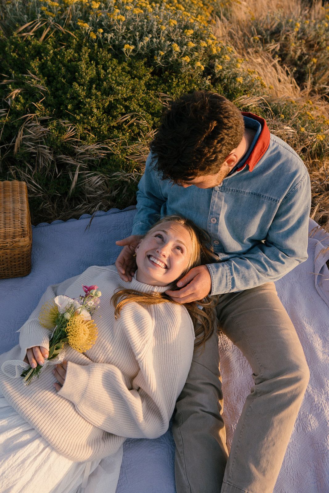 A couple enjoys a romantic picnic on a white blanket with wildflowers at sunset.