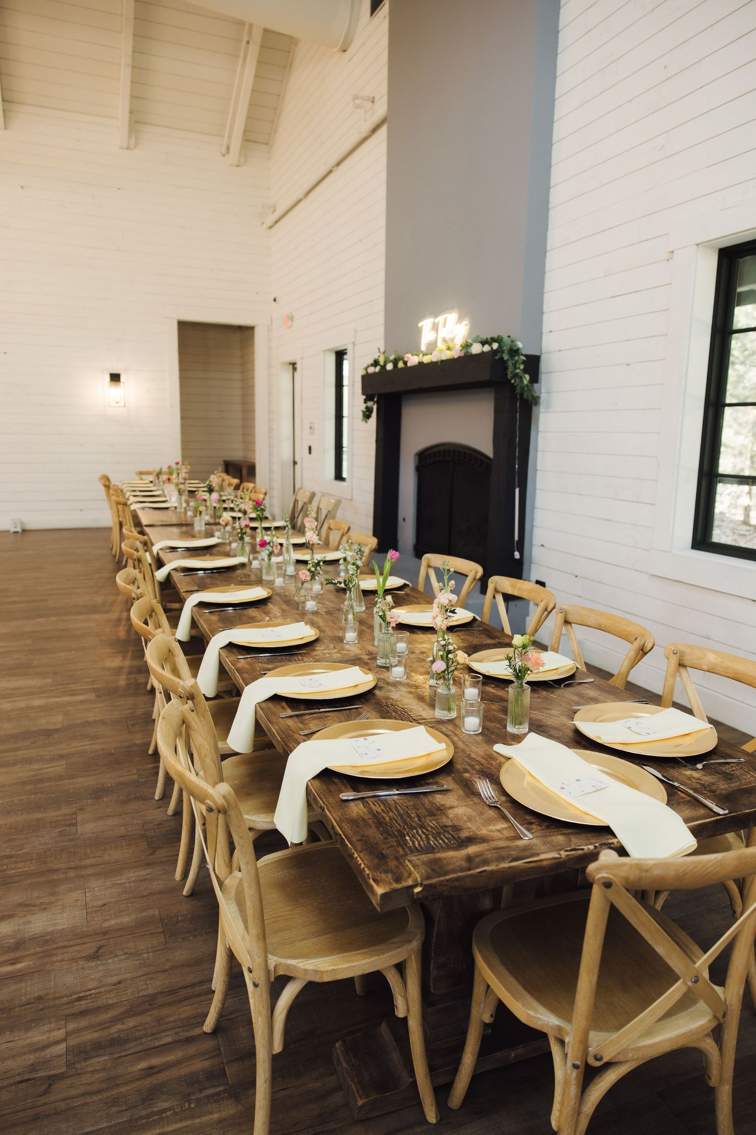 Rustic farmhouse dining table with wooden chairs and elegant table settings in a bright white-walled room.