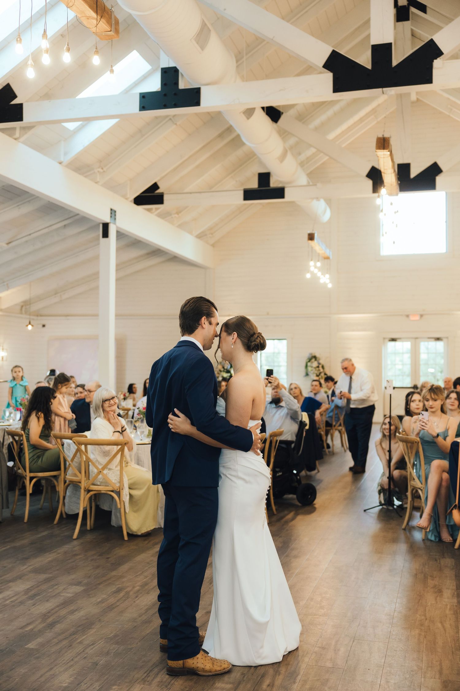 Wedding reception venue with vaulted white ceiling, wood floors and guests dancing in an open hall.