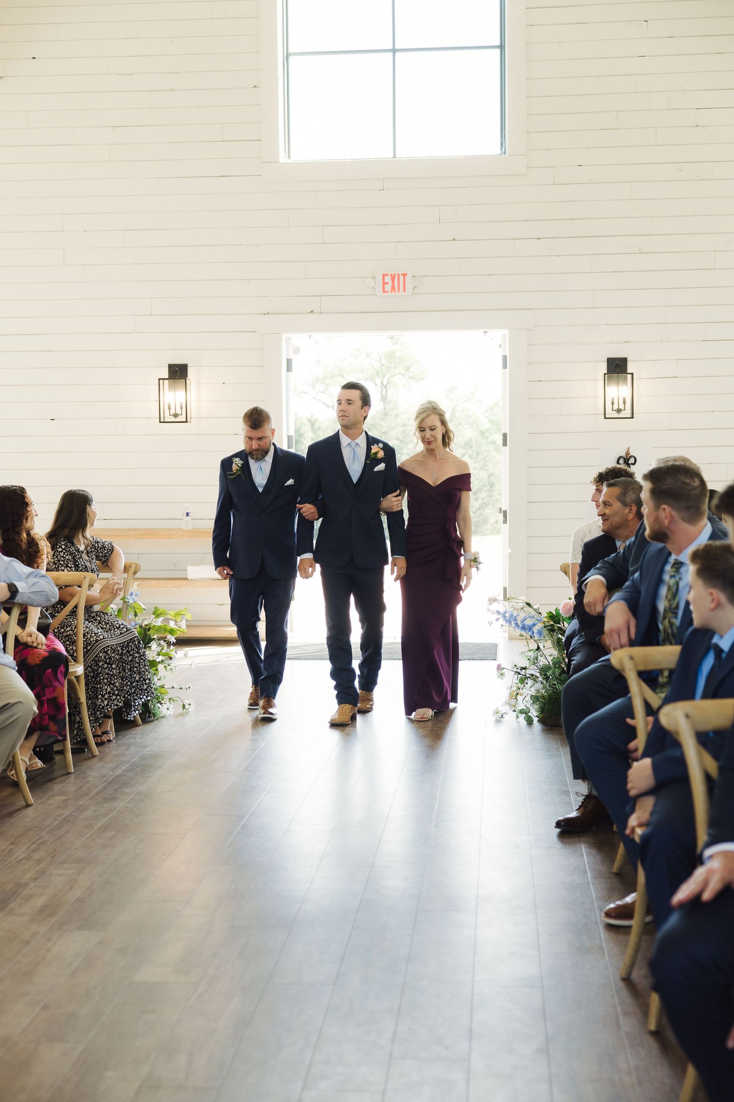 Three attendants walk down an aisle lined with guests and floral arrangements in an elegant white indoor venue.