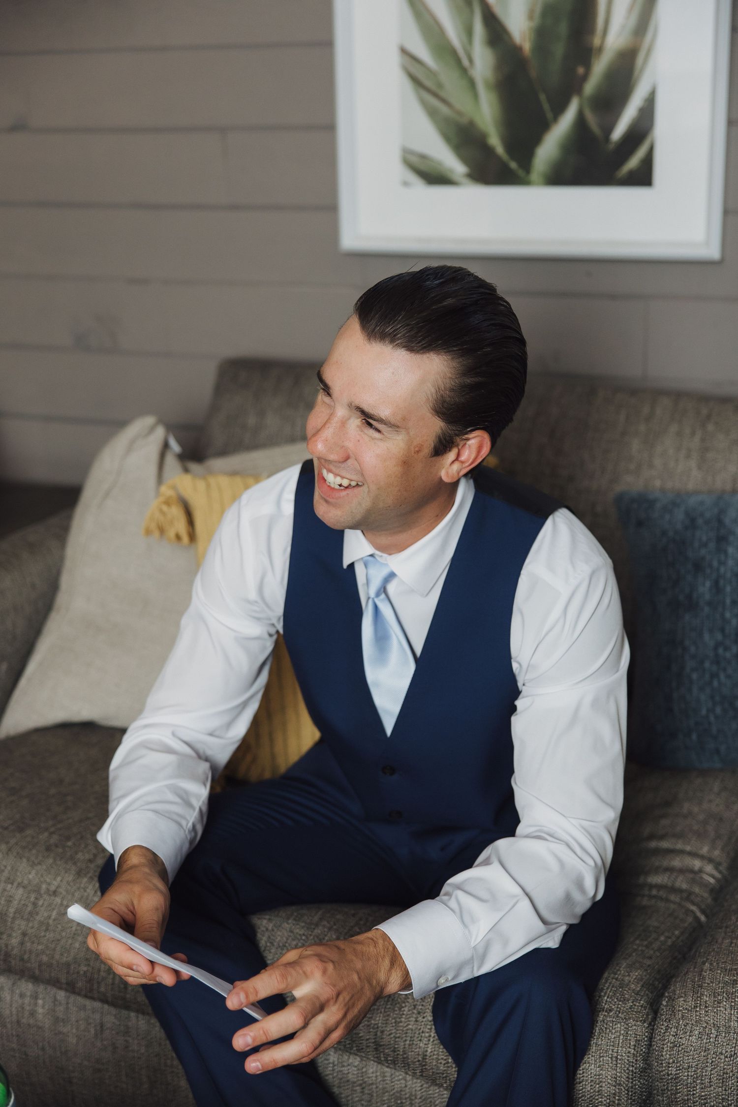 Business professional in navy vest relaxes on grey couch while checking mobile phone during casual moment.