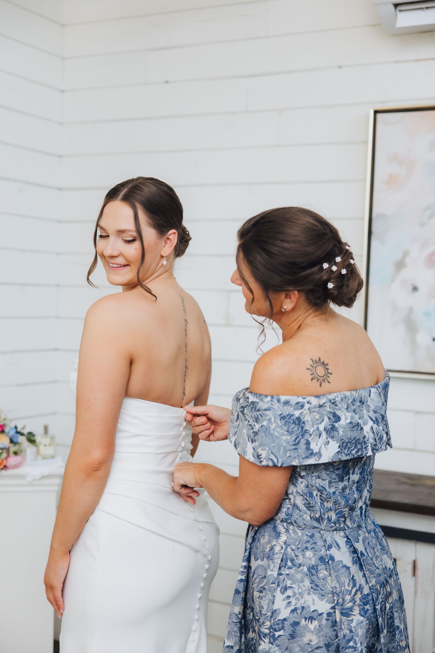 A bride in a white wedding dress gets help with finishing touches while standing next to someone in a blue floral dress.