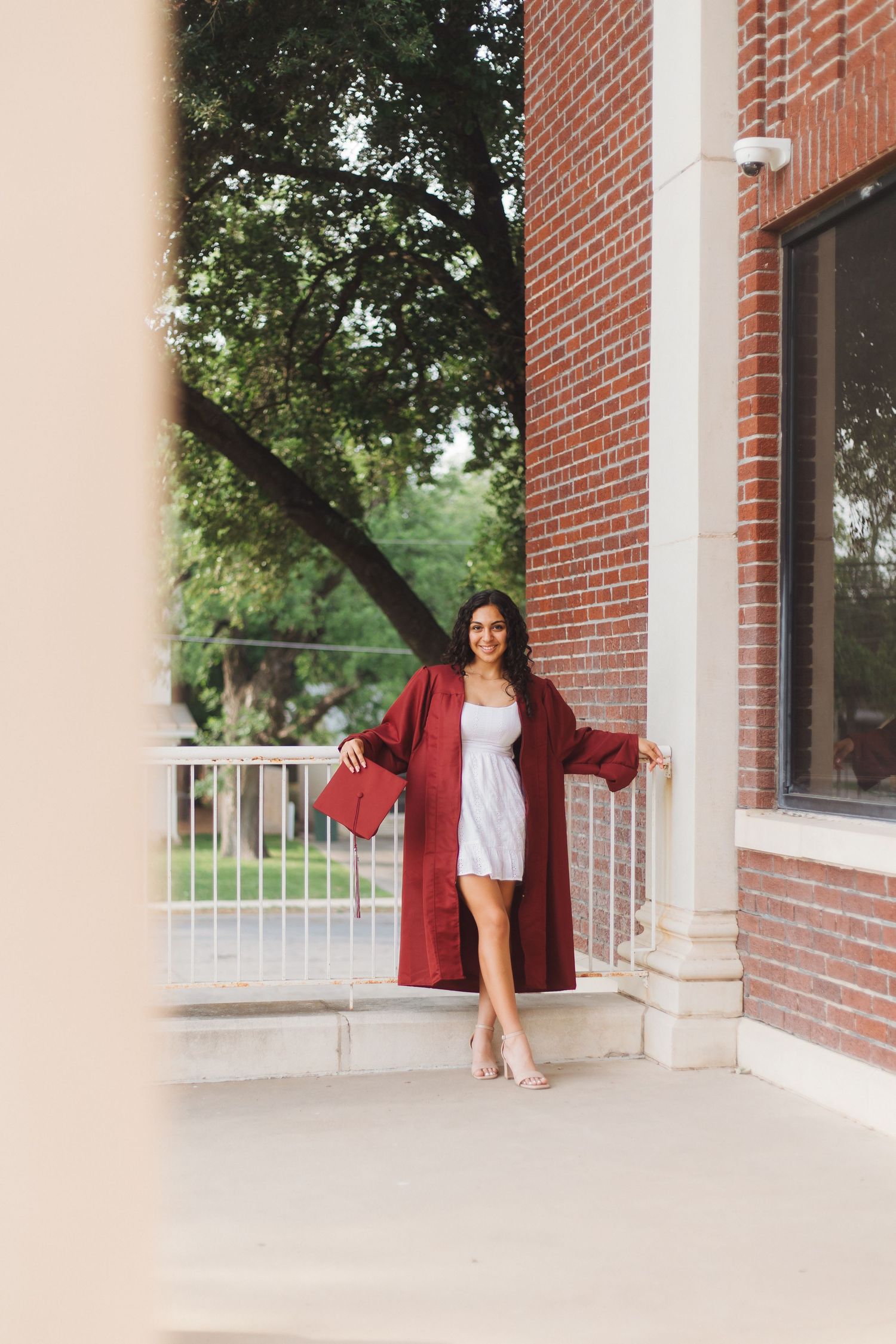A person stands on a brick building porch wearing a long red coat over a white dress and holding a red handbag.