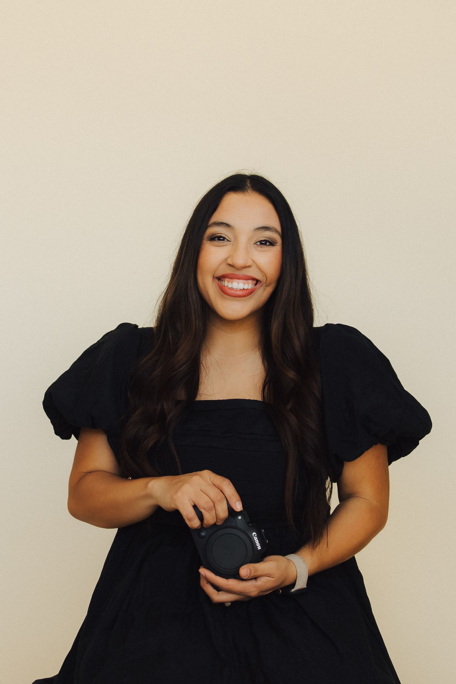 Professional portrait of someone wearing a black puff sleeve dress holding a camera against a beige background.