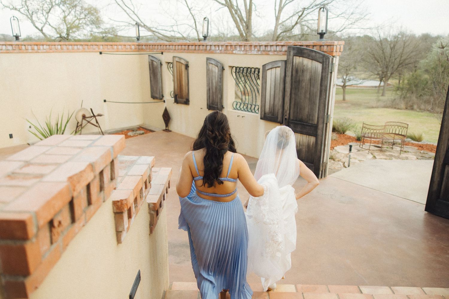 A person in a blue dress and white veil walking up outdoor stairs towards a wooden door on a patio.