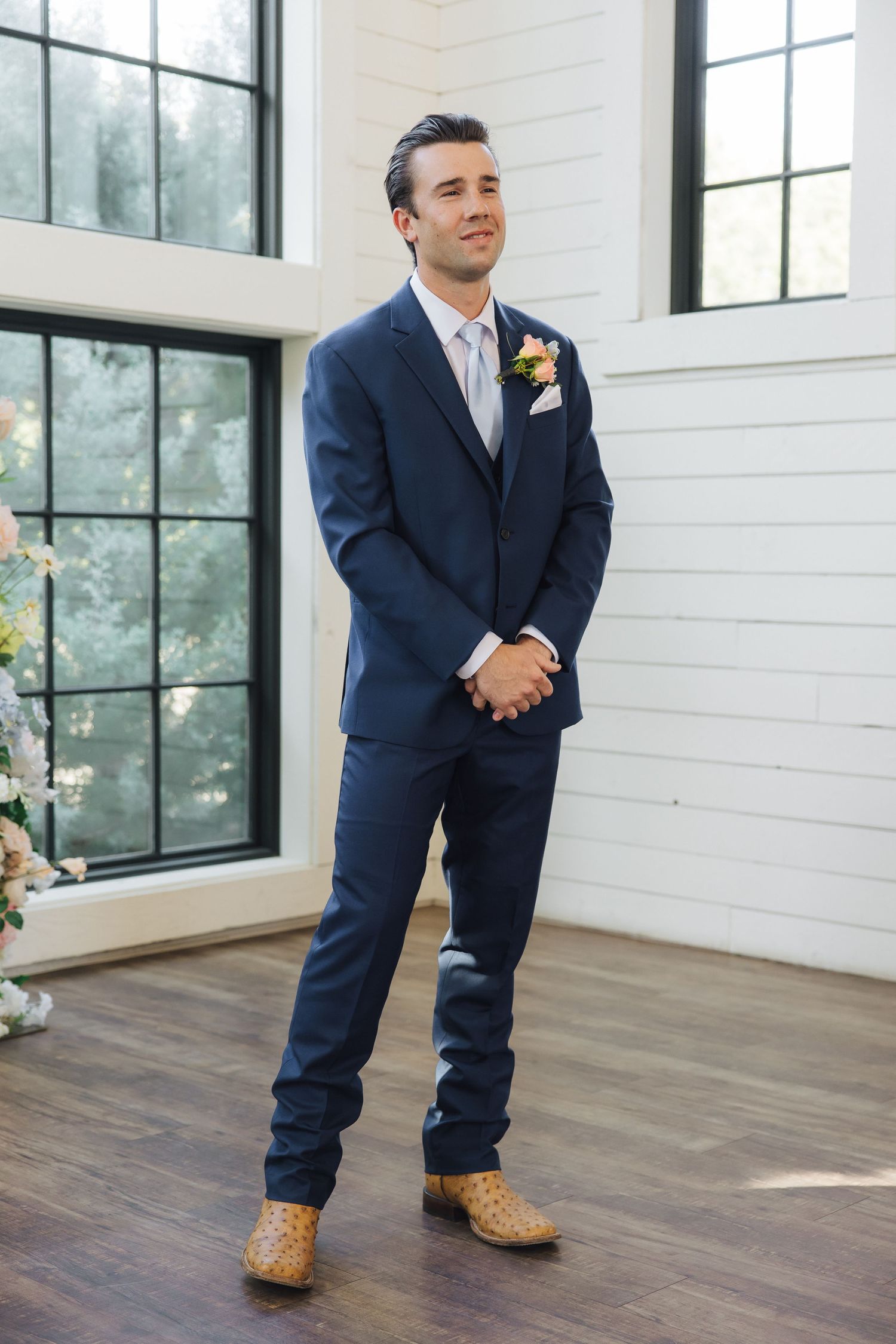 Navy suited groomsman photographed in a sunlit room with white walls and hardwood floors.