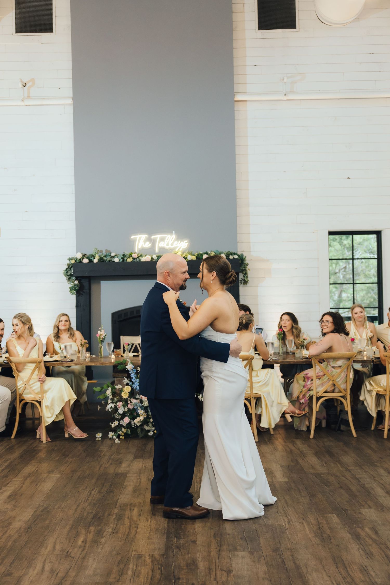 Couples dancing together on a hardwood floor during a wedding reception with white walls and seated guests.