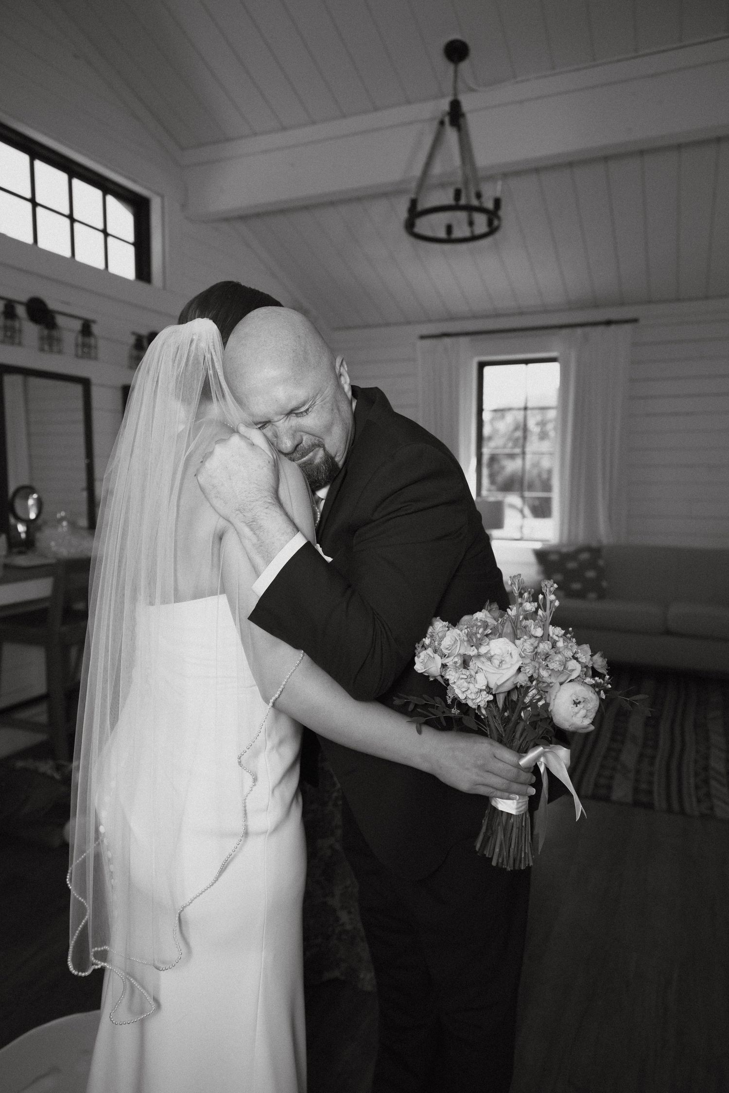 A tender wedding moment captured in black and white with a bridal bouquet and flowing veil in a sunlit room.