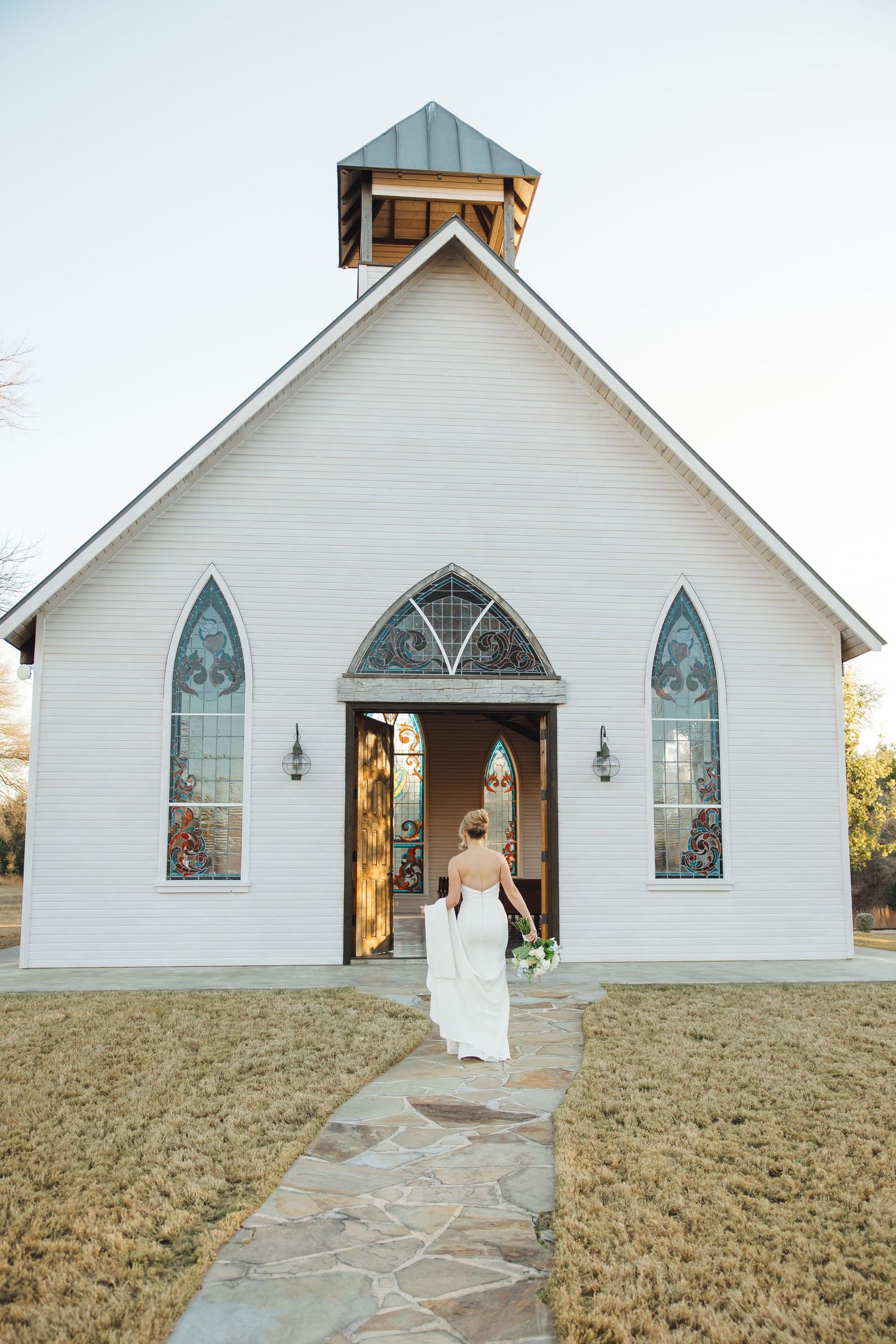 A charming white country chapel with stained glass windows and a stone path leading to open wooden doors.