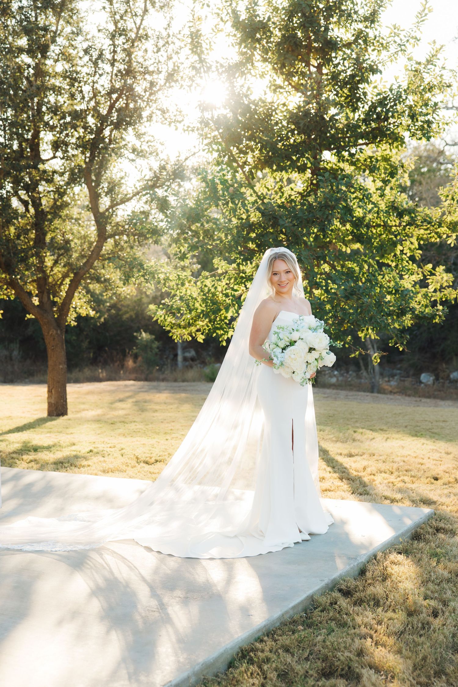 A bride in an elegant white wedding gown and veil holds a bouquet while standing in a sunlit garden setting.