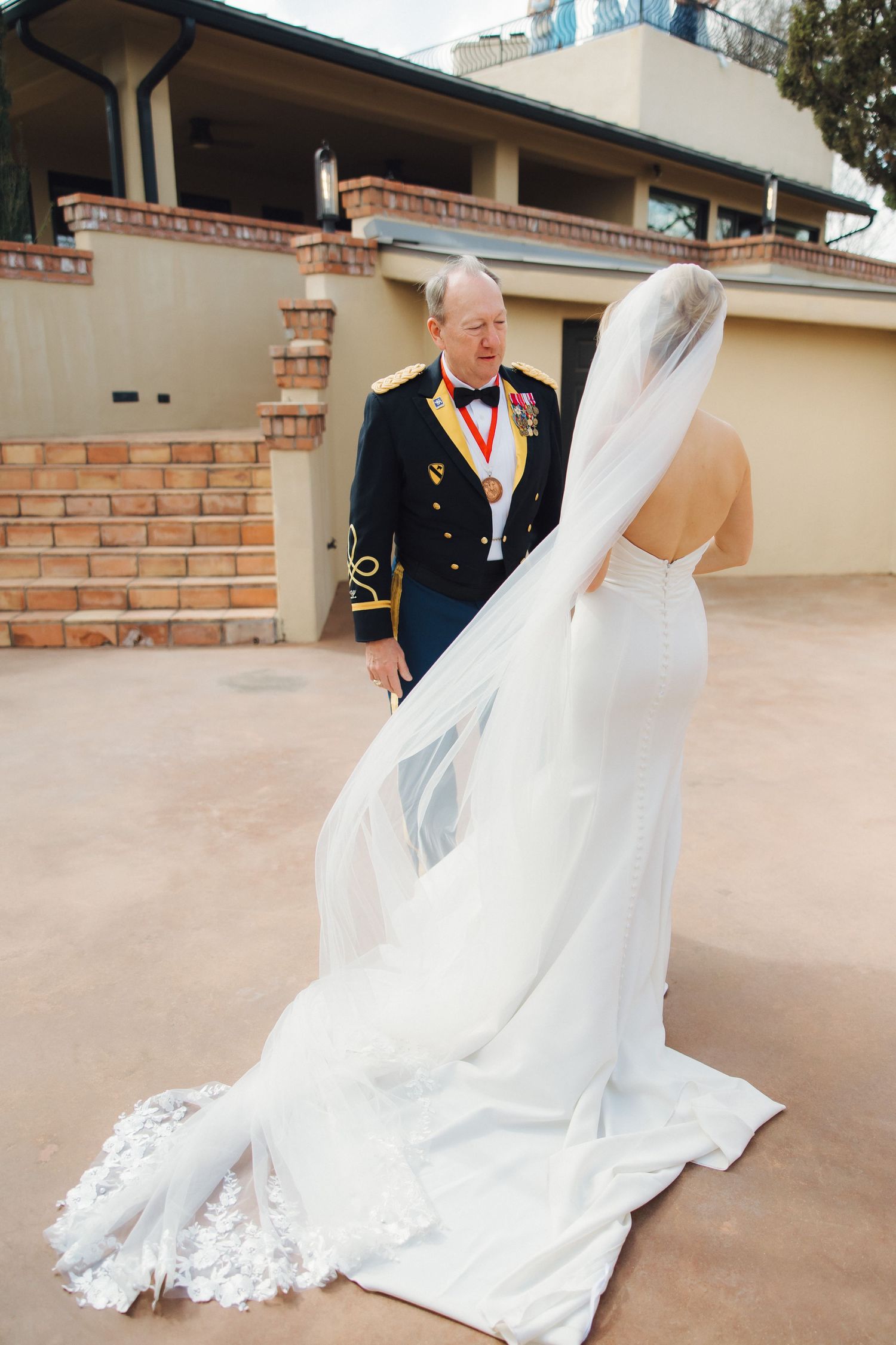 A bride in a flowing white wedding dress with long veil stands with someone in military dress uniform outside a building.