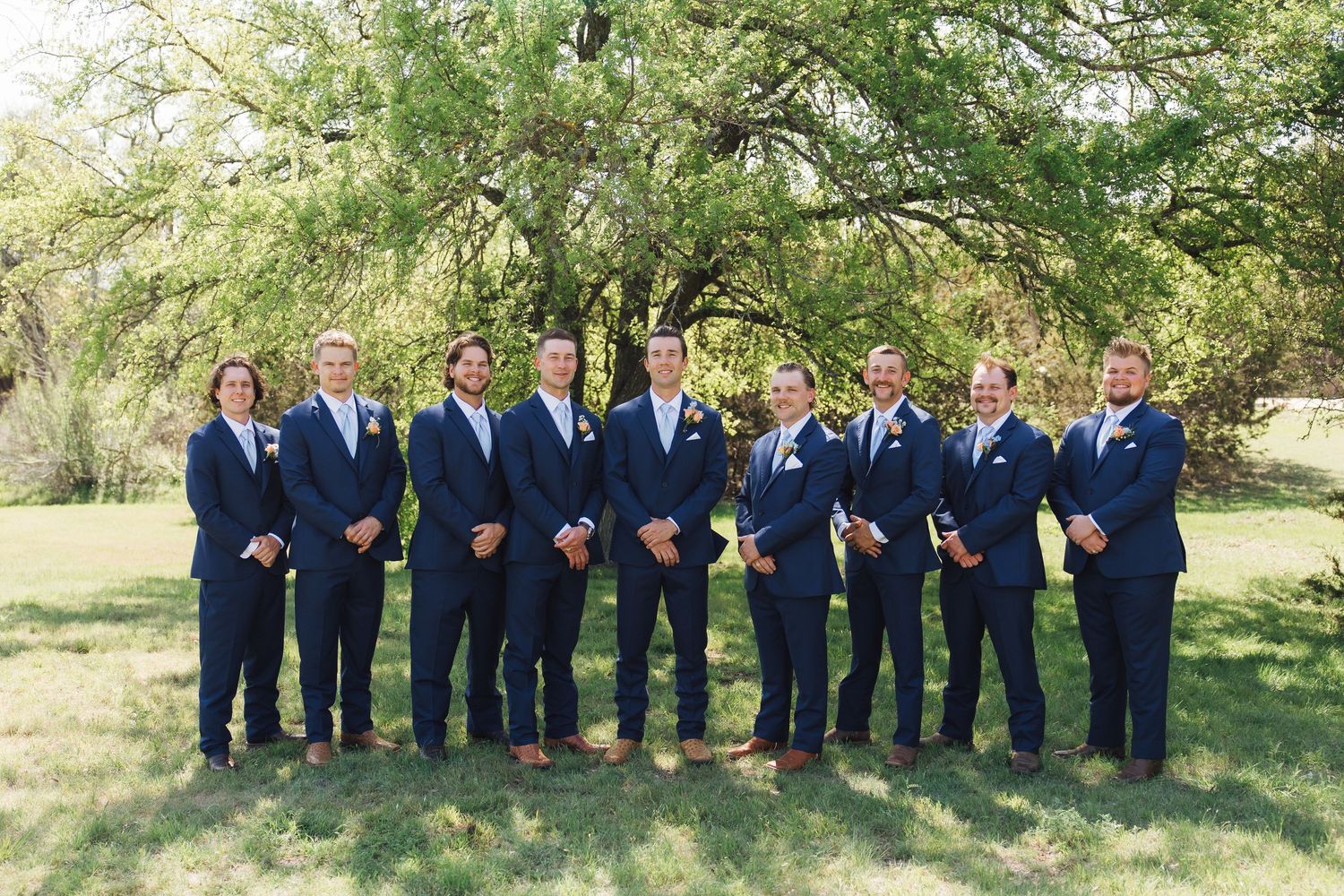 A wedding party wearing matching navy blue suits stands in a line outdoors with green trees in the background.