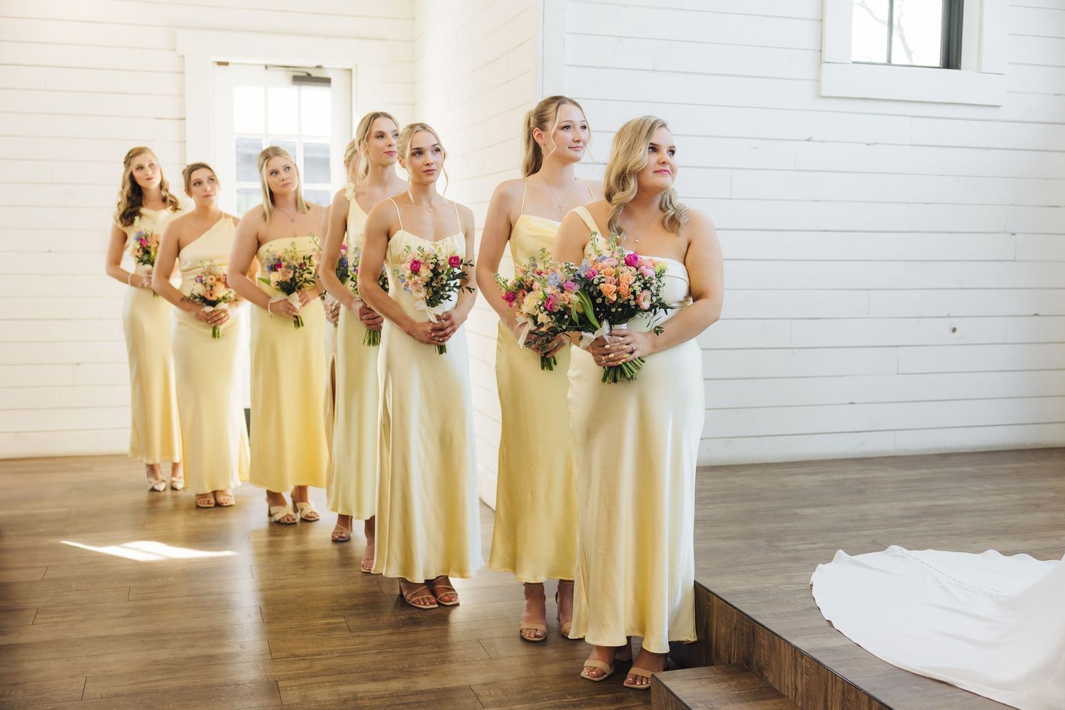 A row of bridesmaids in pale yellow dresses holding floral bouquets stand together in a bright indoor venue.