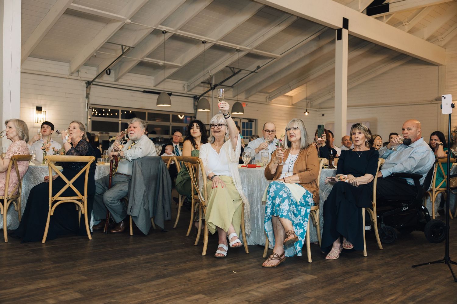 Wedding guests seated at round tables in a bright reception hall with wooden floors and white ceiling beams.