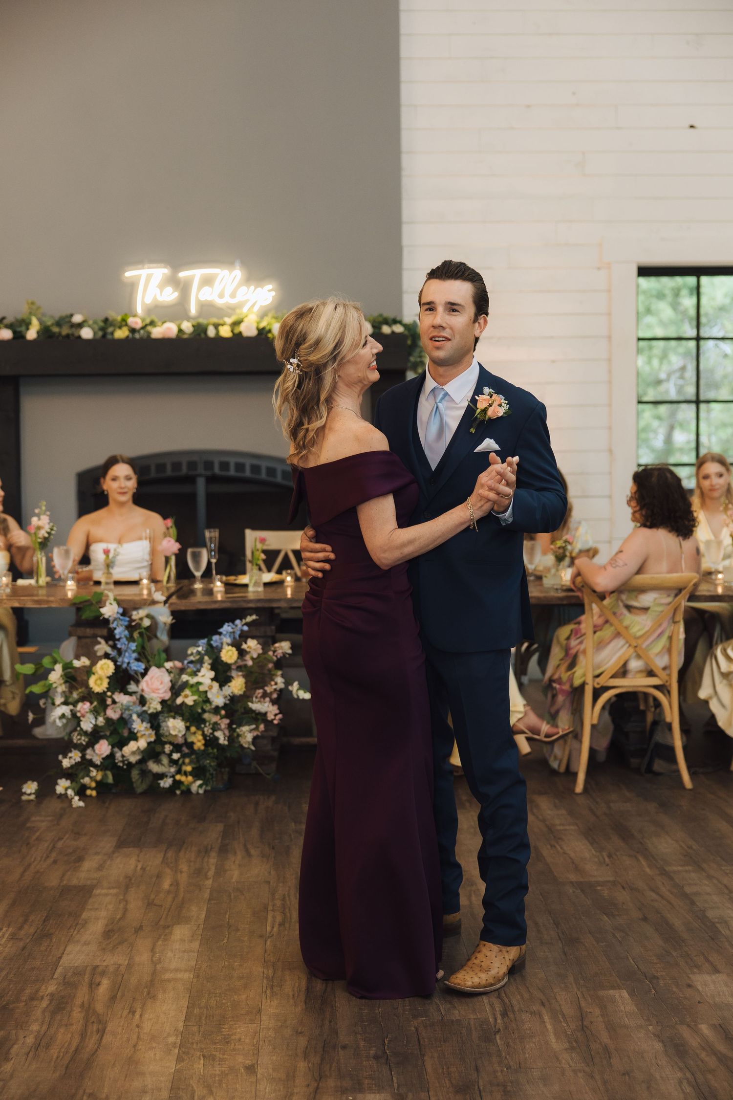 Wedding guests dancing on a wooden floor with white brick walls and decorative lighting in the background.