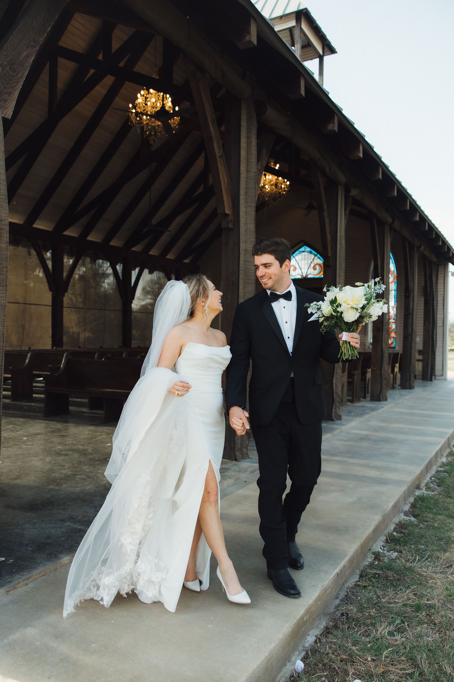 A couple in elegant wedding attire walking together outside a rustic wooden pavilion with hanging chandeliers.