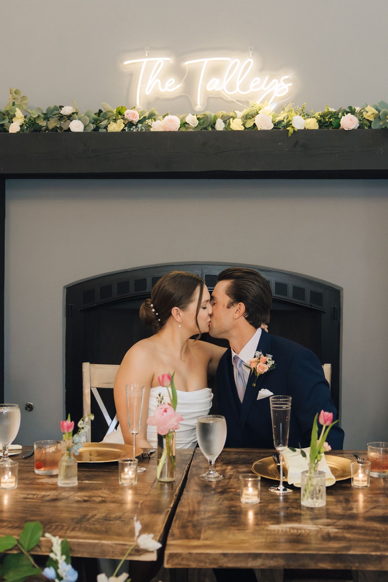Neon sign reading 'The Tallens' illuminates a rustic sweetheart table decorated with flowers and glassware.