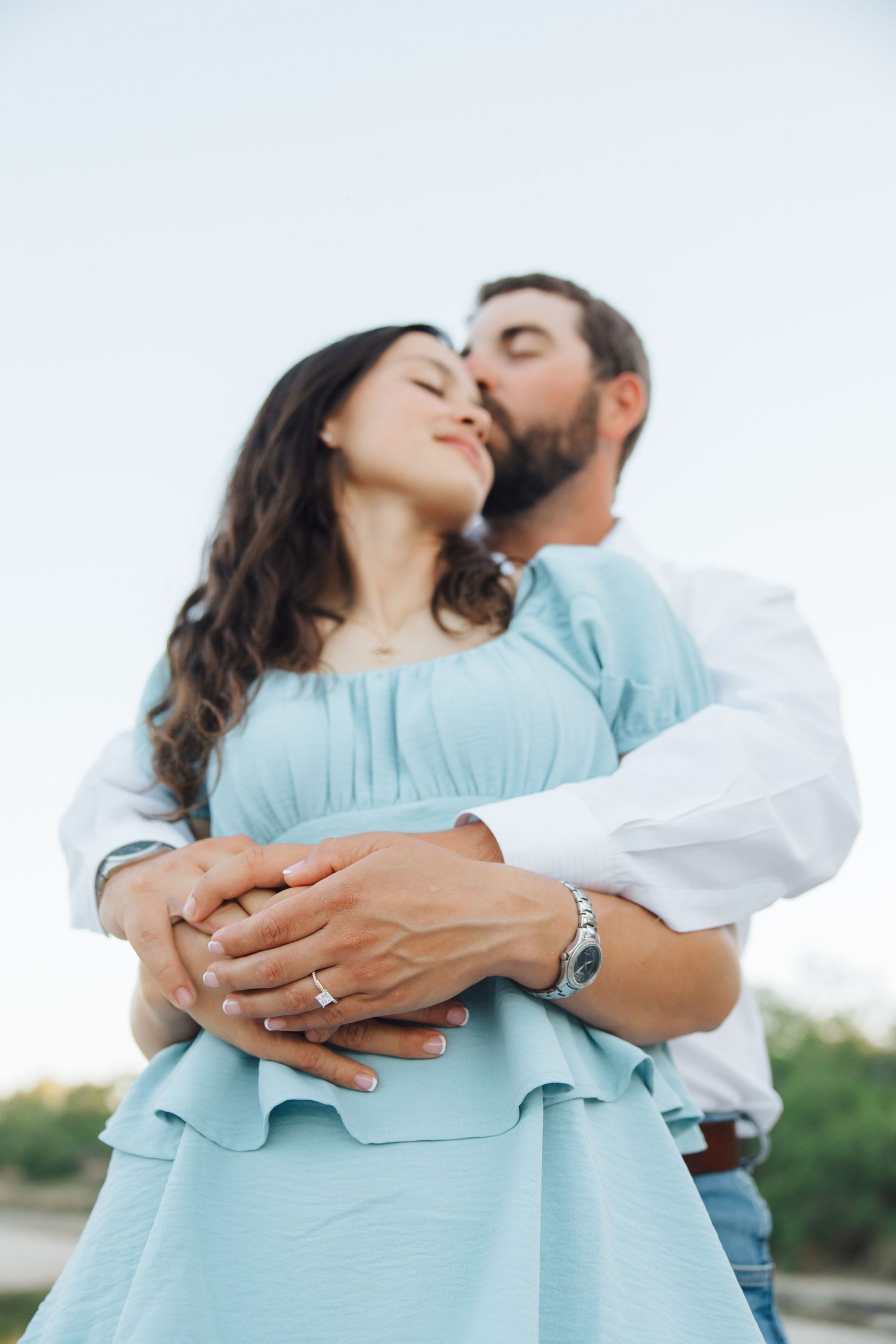 A romantic couple in light blue and white clothing embrace against a bright outdoor backdrop.