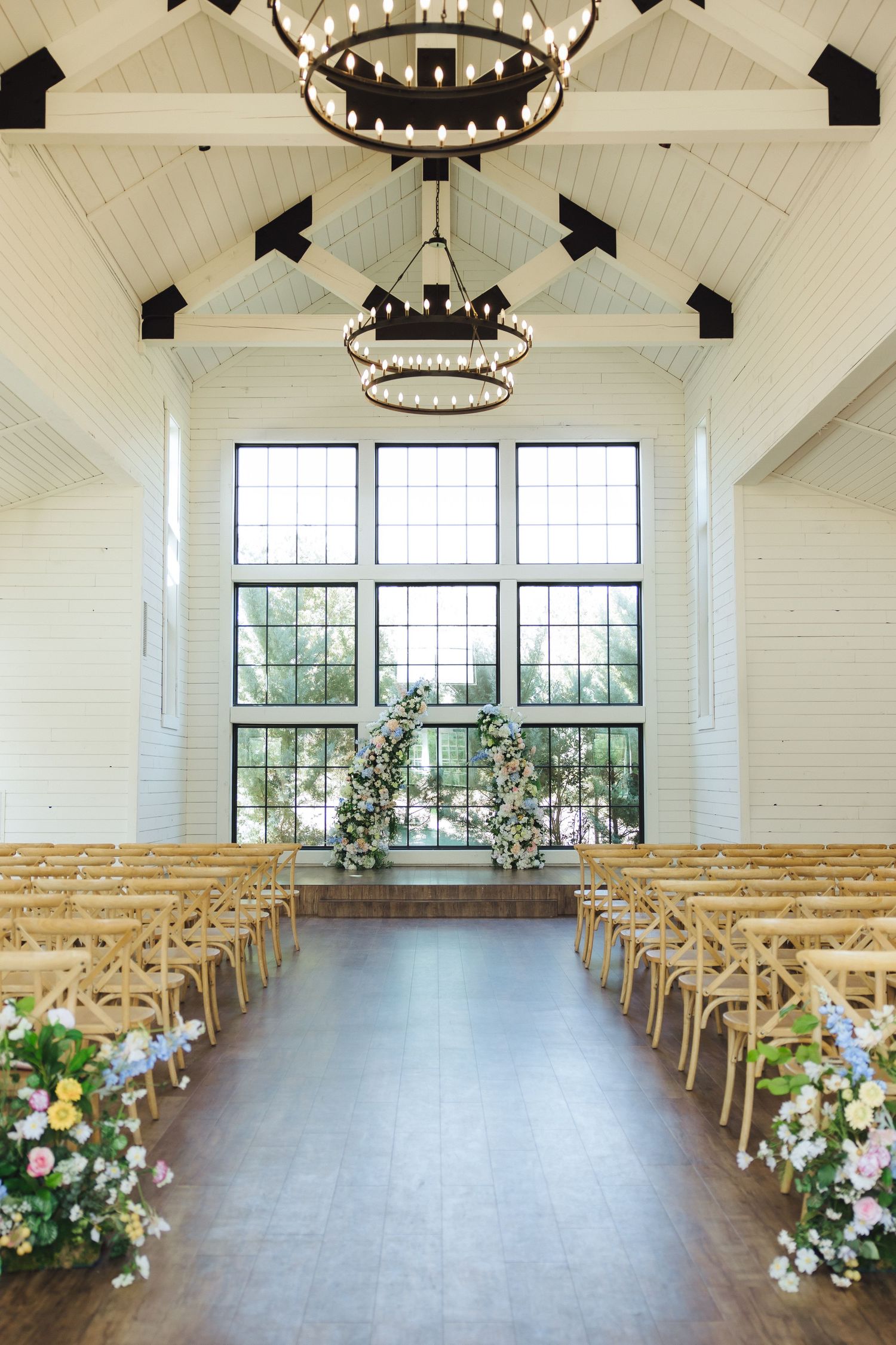 Elegant wedding ceremony venue with rows of wooden chairs, white walls, chandeliers and large windows overlooking greenery.