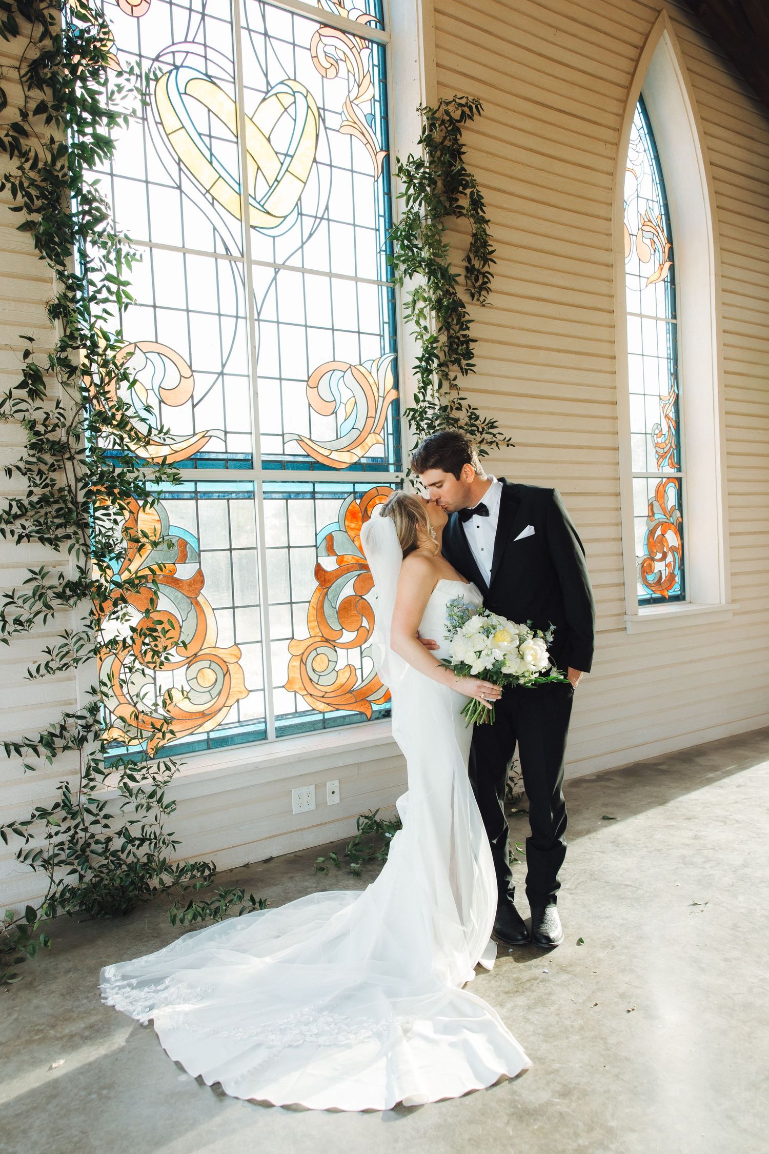 A couple shares an intimate moment in front of a stunning stained glass window with trailing greenery in a vintage chapel.