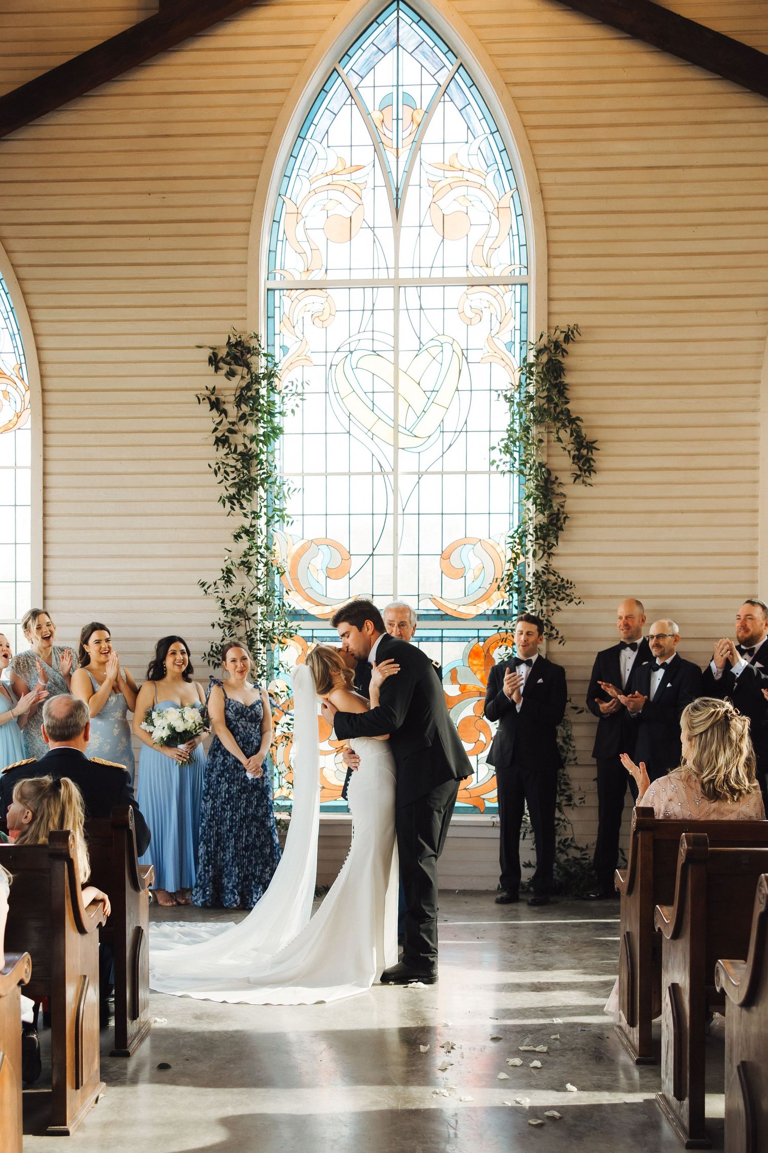 Couple shares a romantic kiss at the altar in a rustic chapel with stained glass windows and greenery decorations.