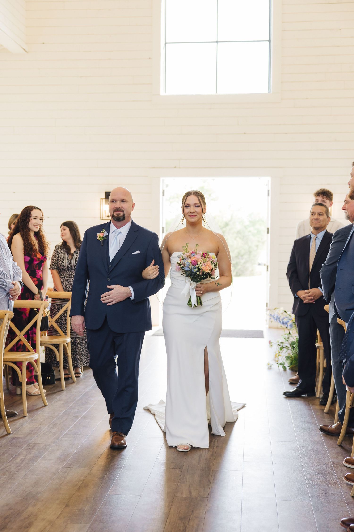 A bride walks down the aisle with an escort at a rustic indoor wedding venue with wooden floors and natural lighting.