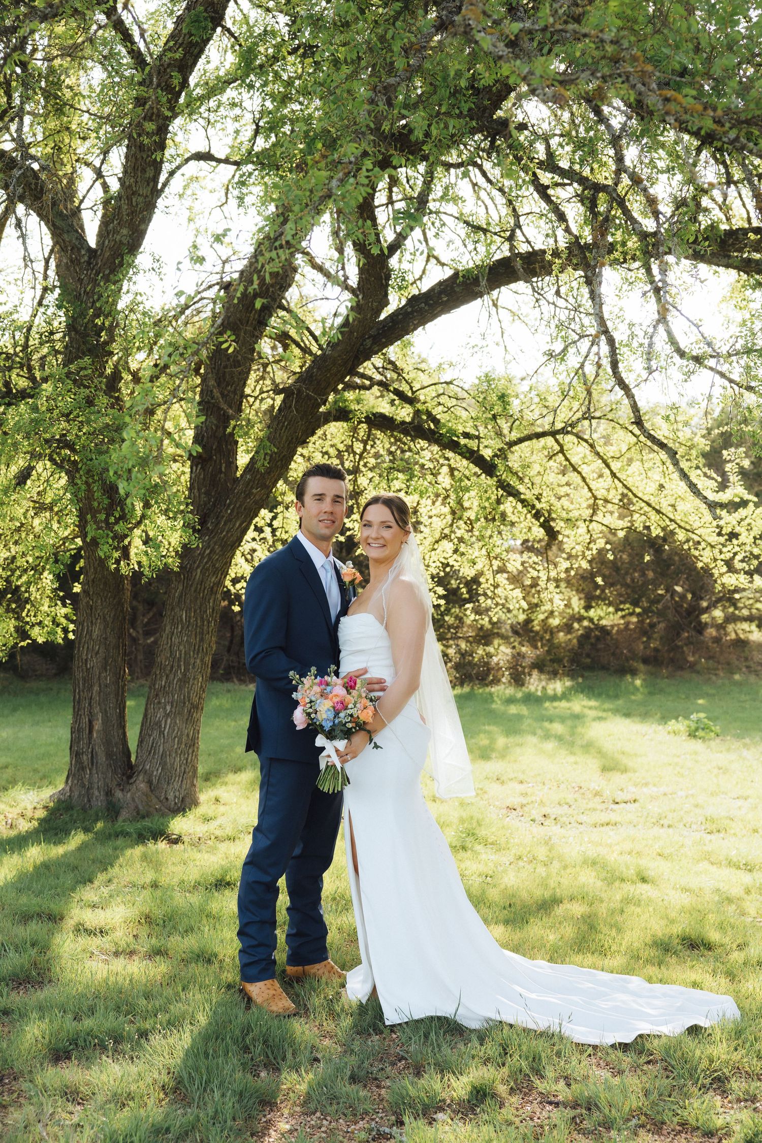 A couple in formal wedding attire poses under a leafy green tree on a sunny day in nature.