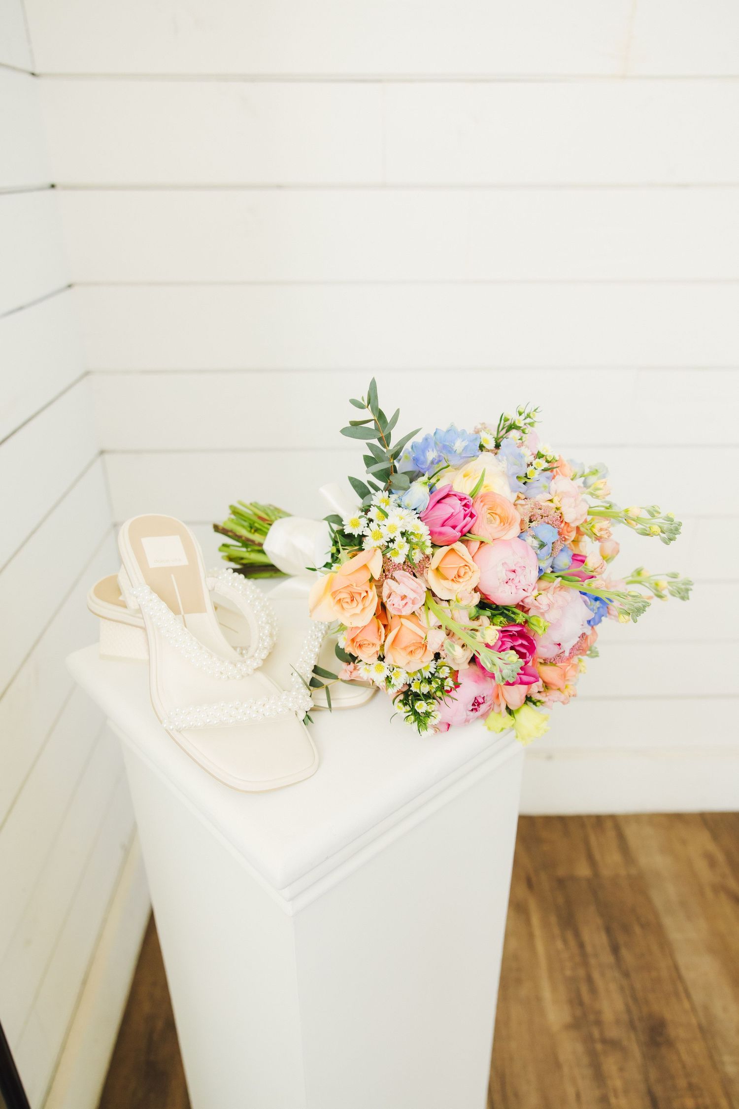 A colorful spring bouquet of mixed flowers sits next to blush wedding sandals on a white pedestal against white walls.