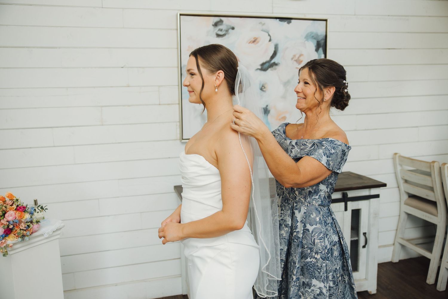 A woman in a blue floral dress helps another woman put on a white wedding gown in a bright indoor setting.