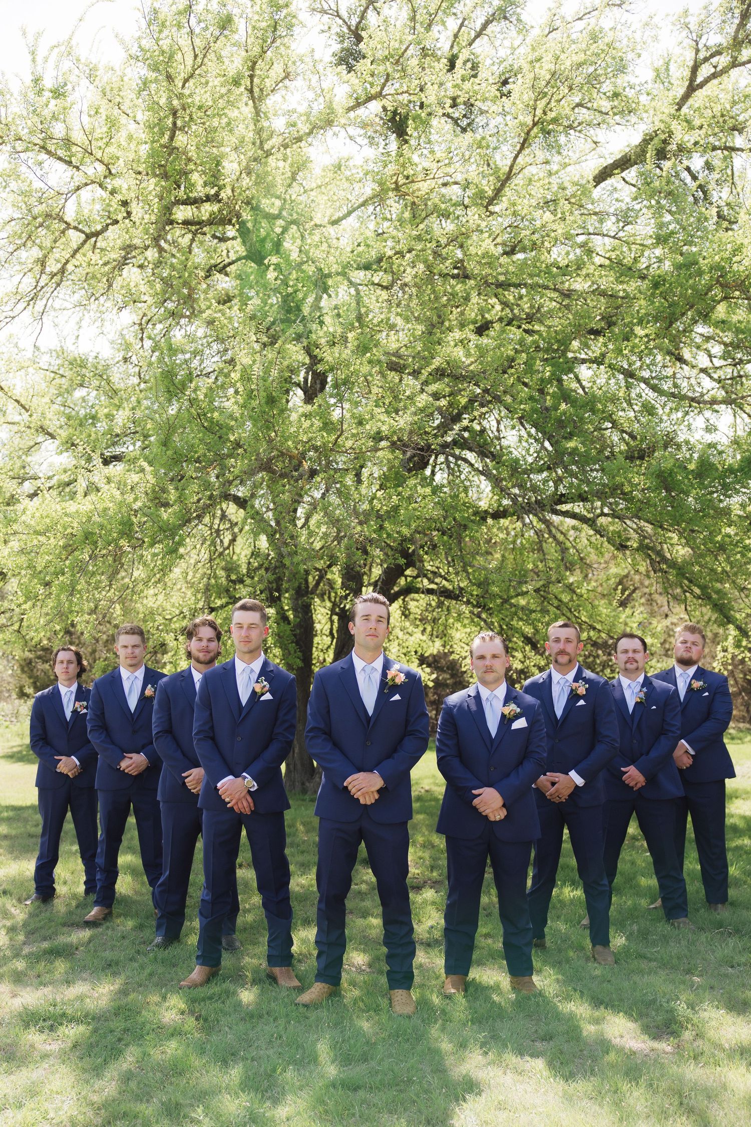 A wedding party in matching navy suits and brown shoes poses outdoors under a large green tree on grass.