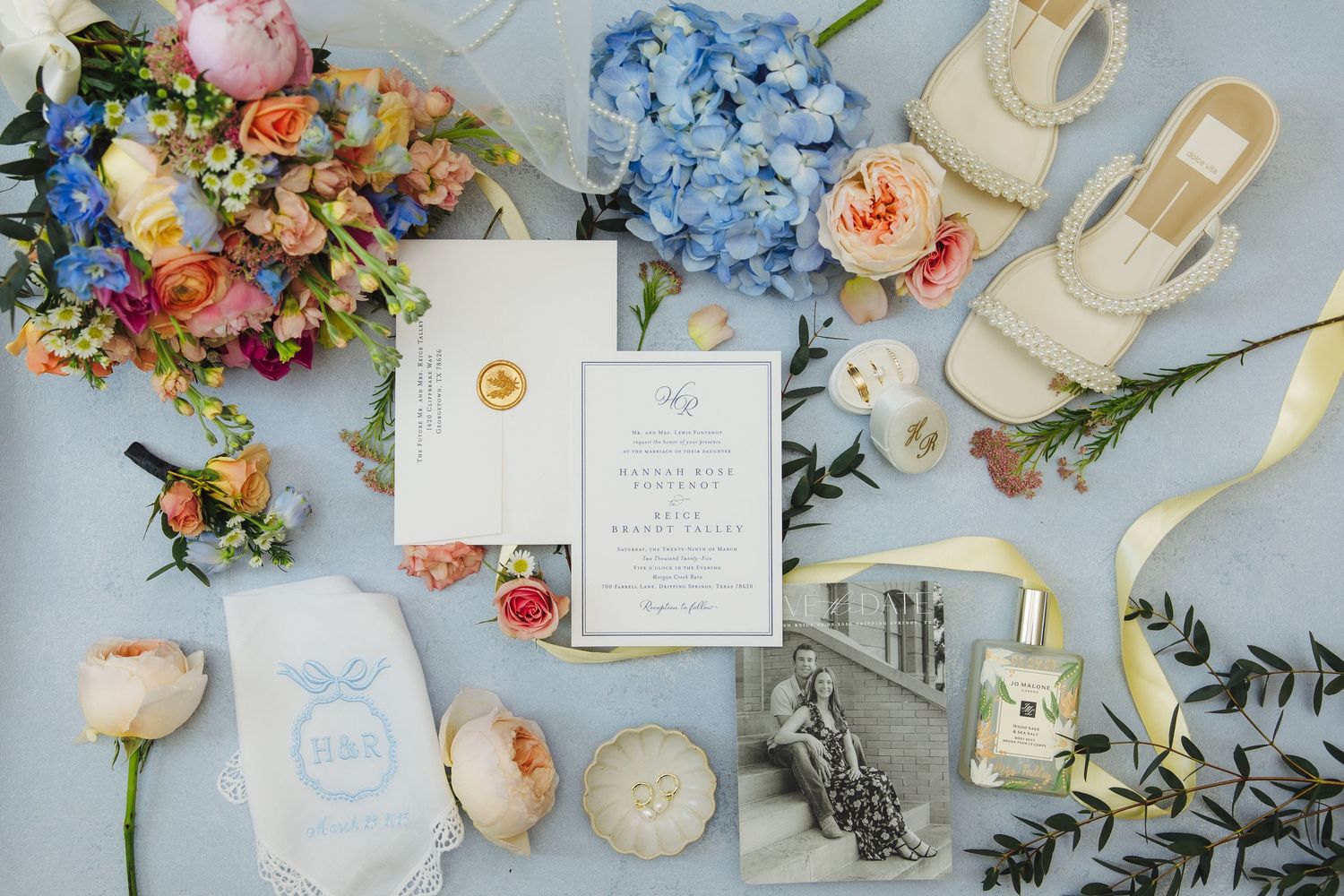 A wedding invitation flat lay with floral arrangements, blue hydrangeas, cream shoes, and perfume bottle on a light surface.