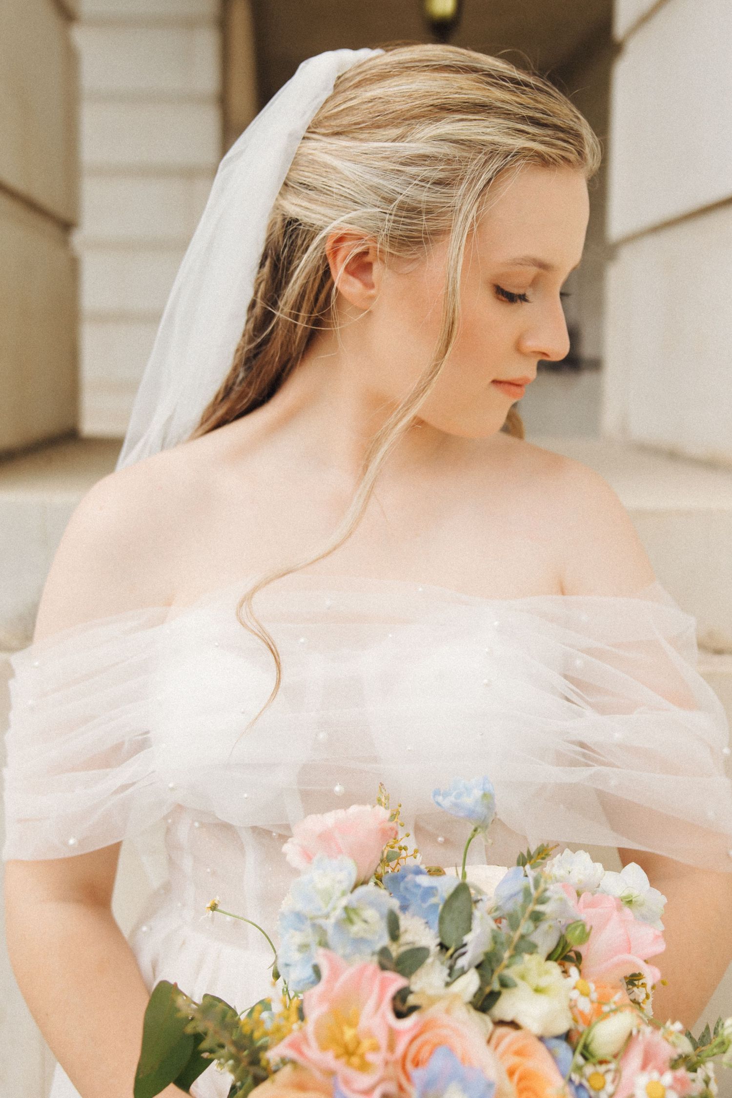 Elegant bride in off-shoulder dress holds pastel bouquet of pink, blue and white flowers with delicate tulle veil.
