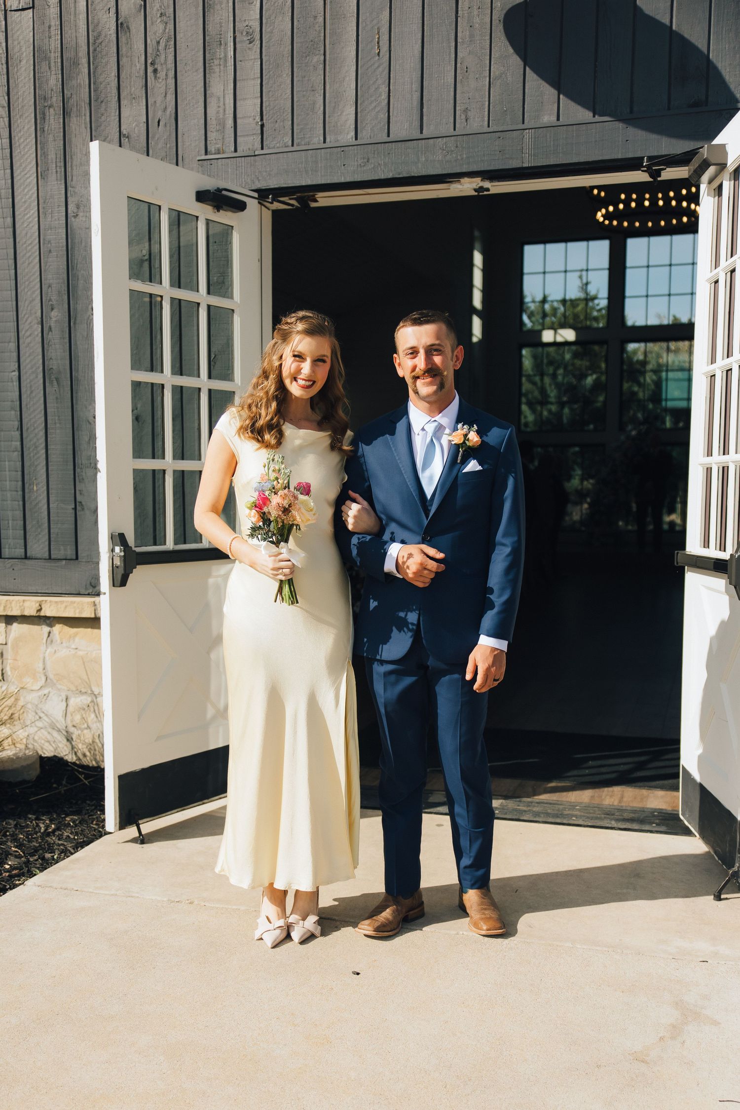 A couple stands in a doorway of a gray barn venue on their wedding day, wearing formal attire and holding a bouquet.