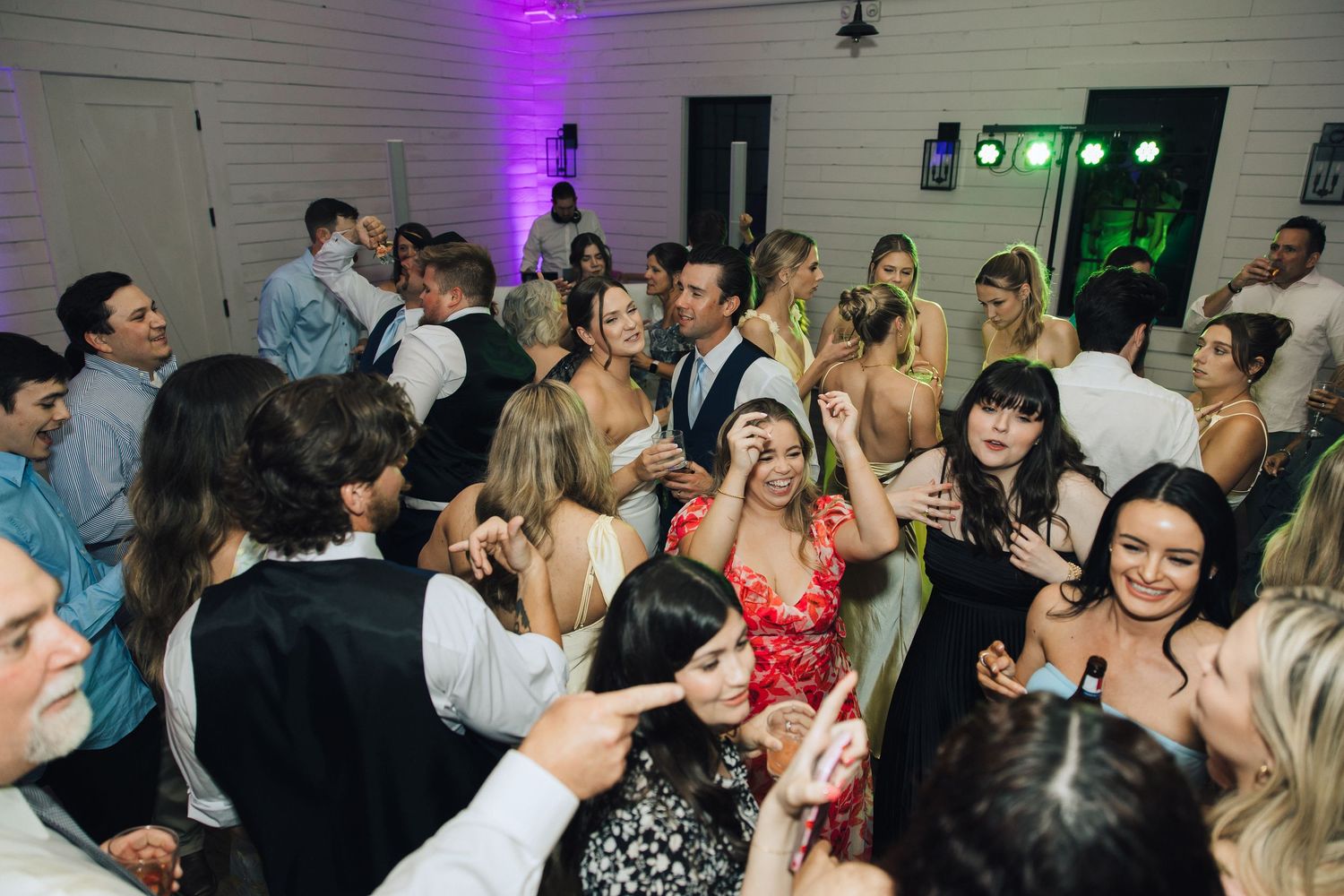 Energetic crowd dancing and celebrating at a wedding reception with purple lighting and white walls.