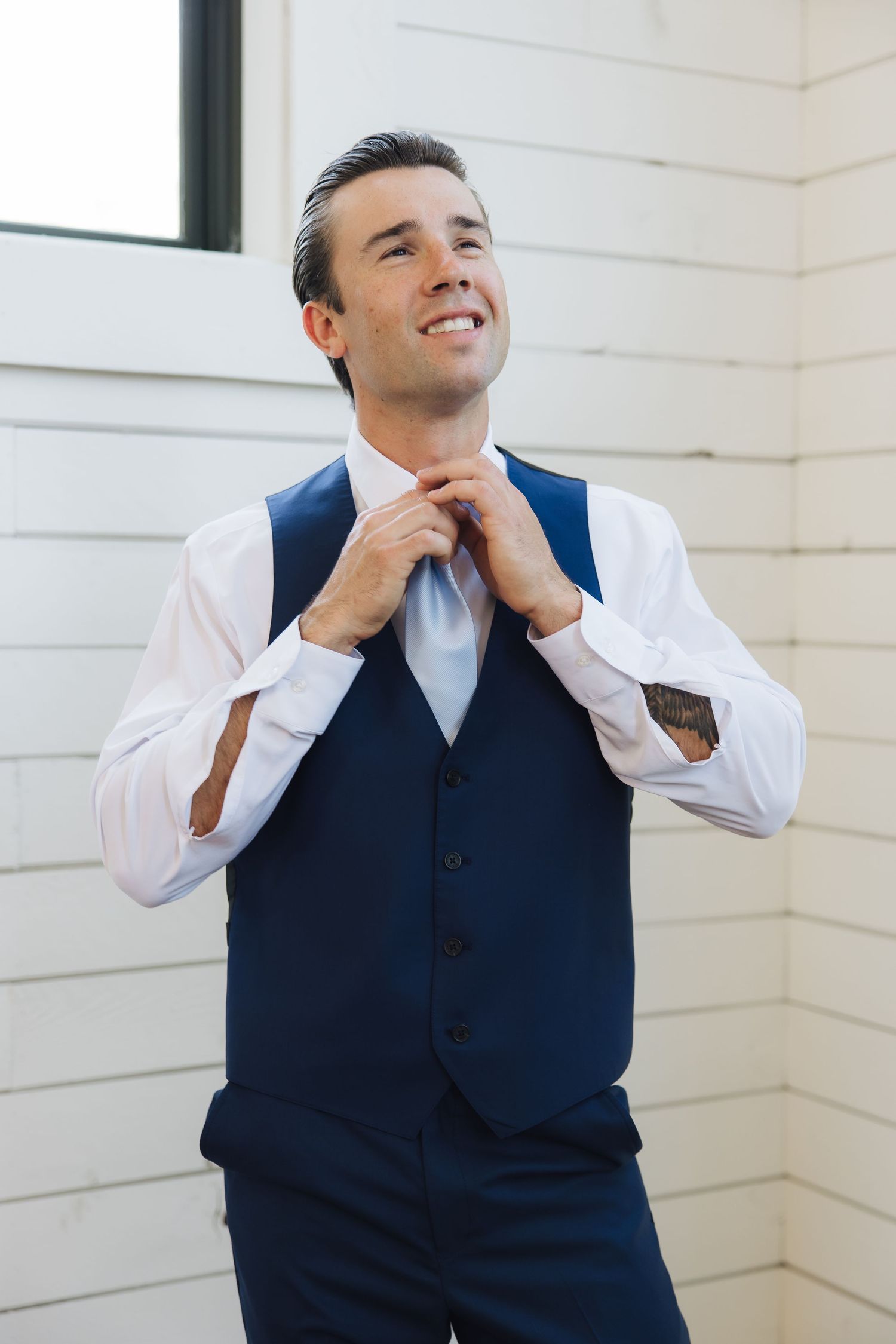 A person in a navy blue vest and white dress shirt adjusts their tie while smiling against a white wall.