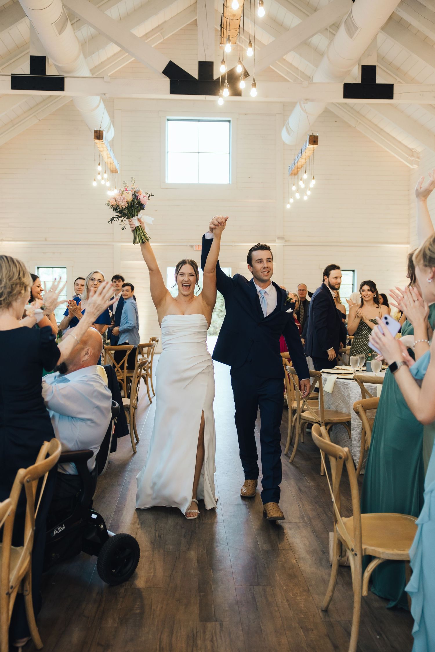 A newly married couple makes their grand entrance into a rustic wedding reception venue with vaulted white ceilings.