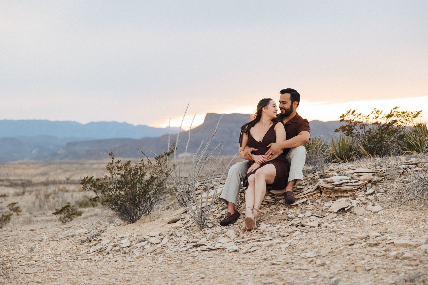 A romantic couple sits on a rocky overlook during sunset in a desert mountain landscape with distant cliffs.