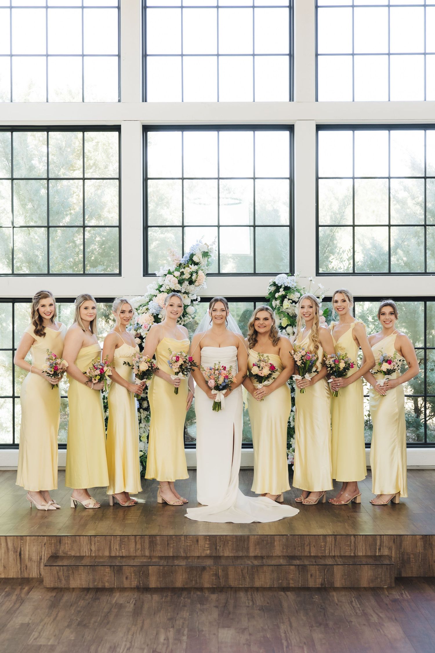 Wedding party in pale yellow dresses poses on rustic wooden platform in bright sunlit venue with large windows.