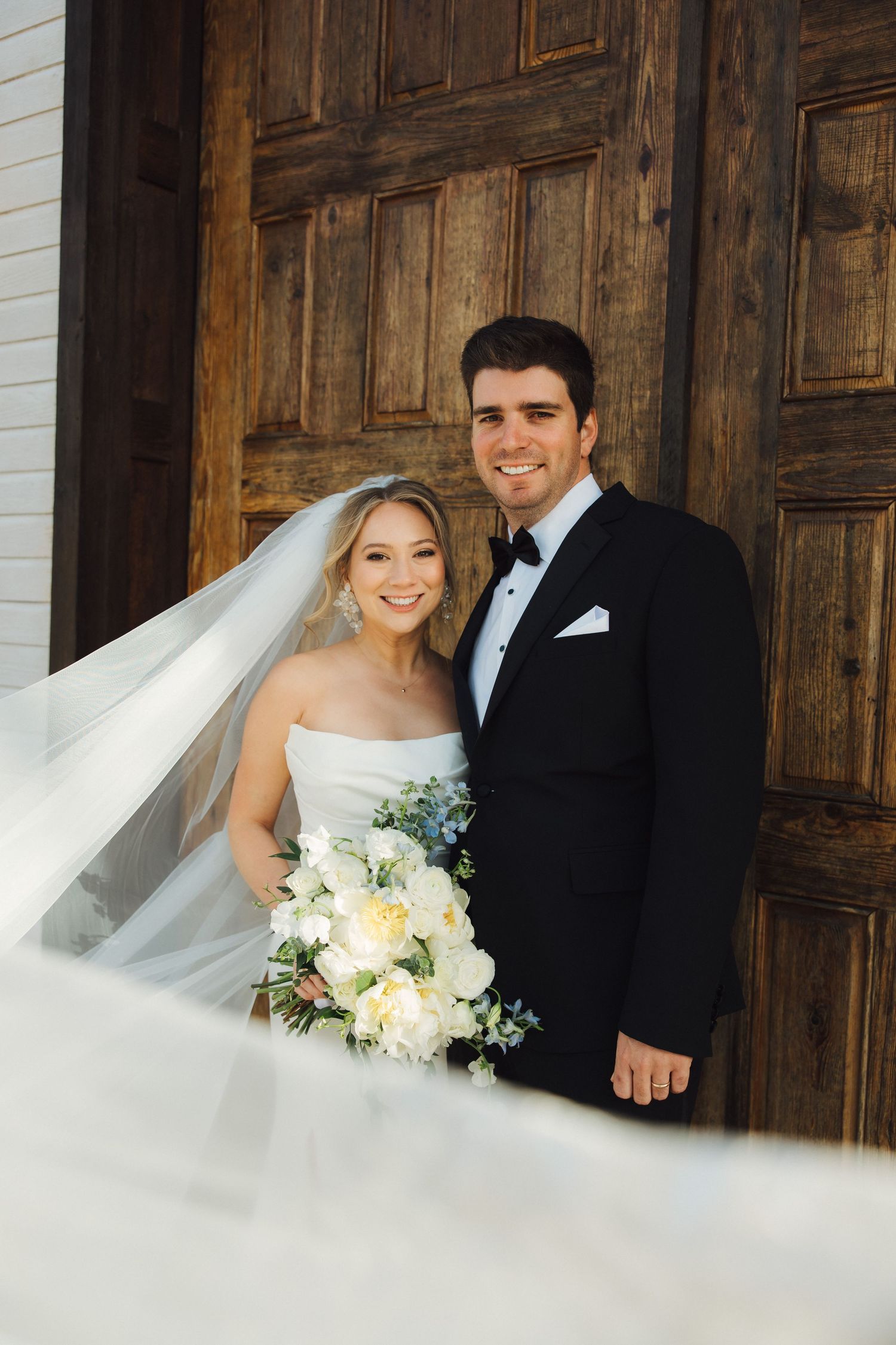 A wedding couple poses in front of a grand wooden door wearing formal attire with a white floral bouquet.