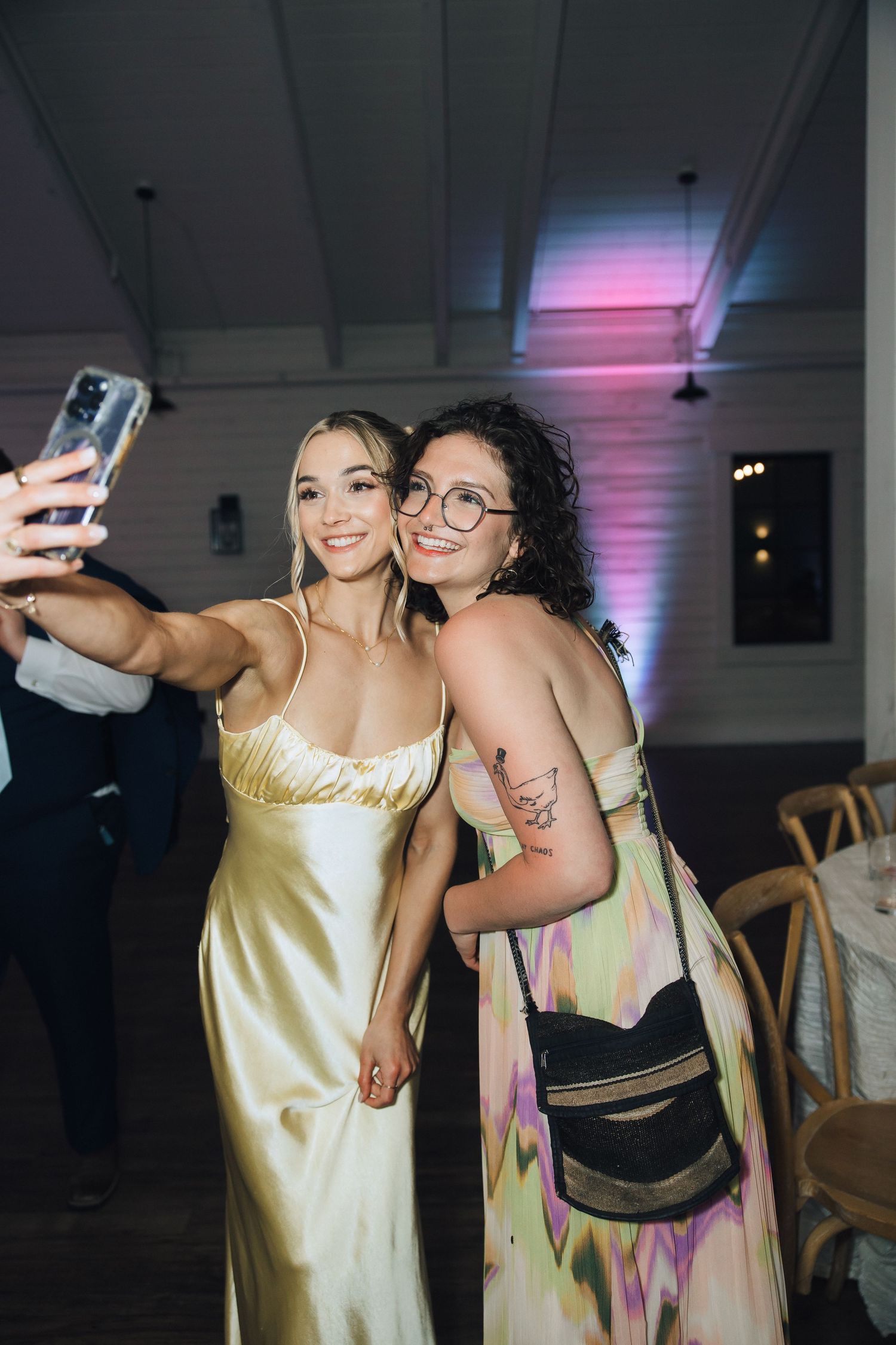 Two friends take a selfie together at an event, one wearing a silky gold dress and another in a patterned pastel outfit.