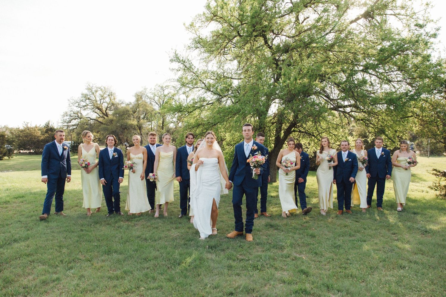 A wedding party stands together on a grassy hillside for outdoor group photos in natural lighting.