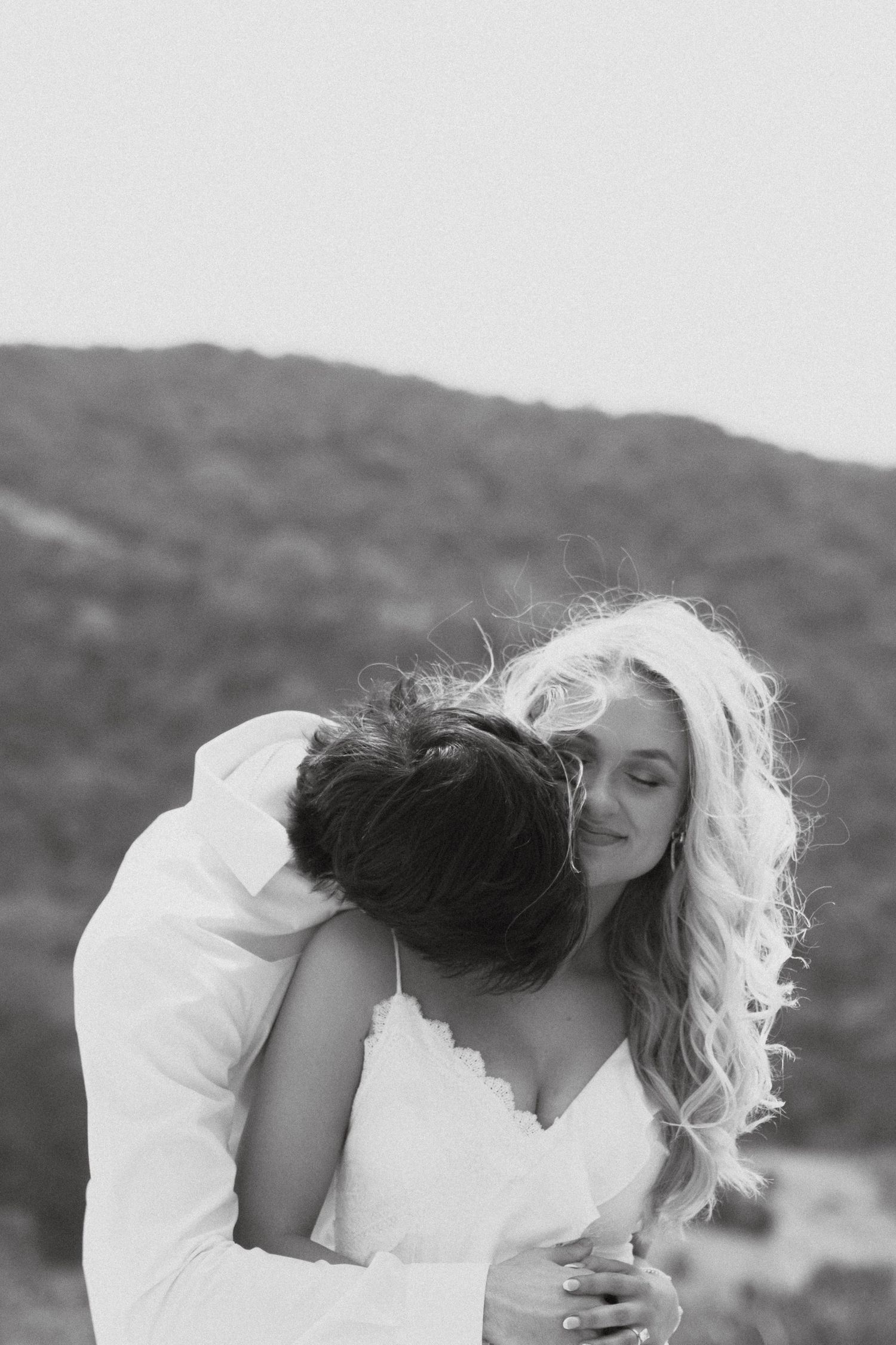 Black and white romantic photo of a couple sharing an intimate embrace outdoors with mountains in the background.