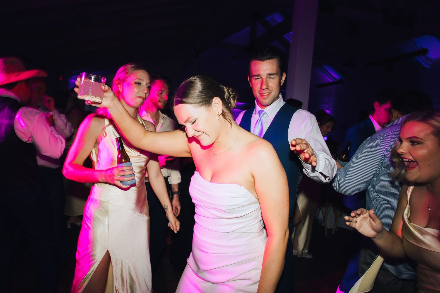 People dance and celebrate at a nightclub with colorful pink and blue lighting creating an energetic party atmosphere.