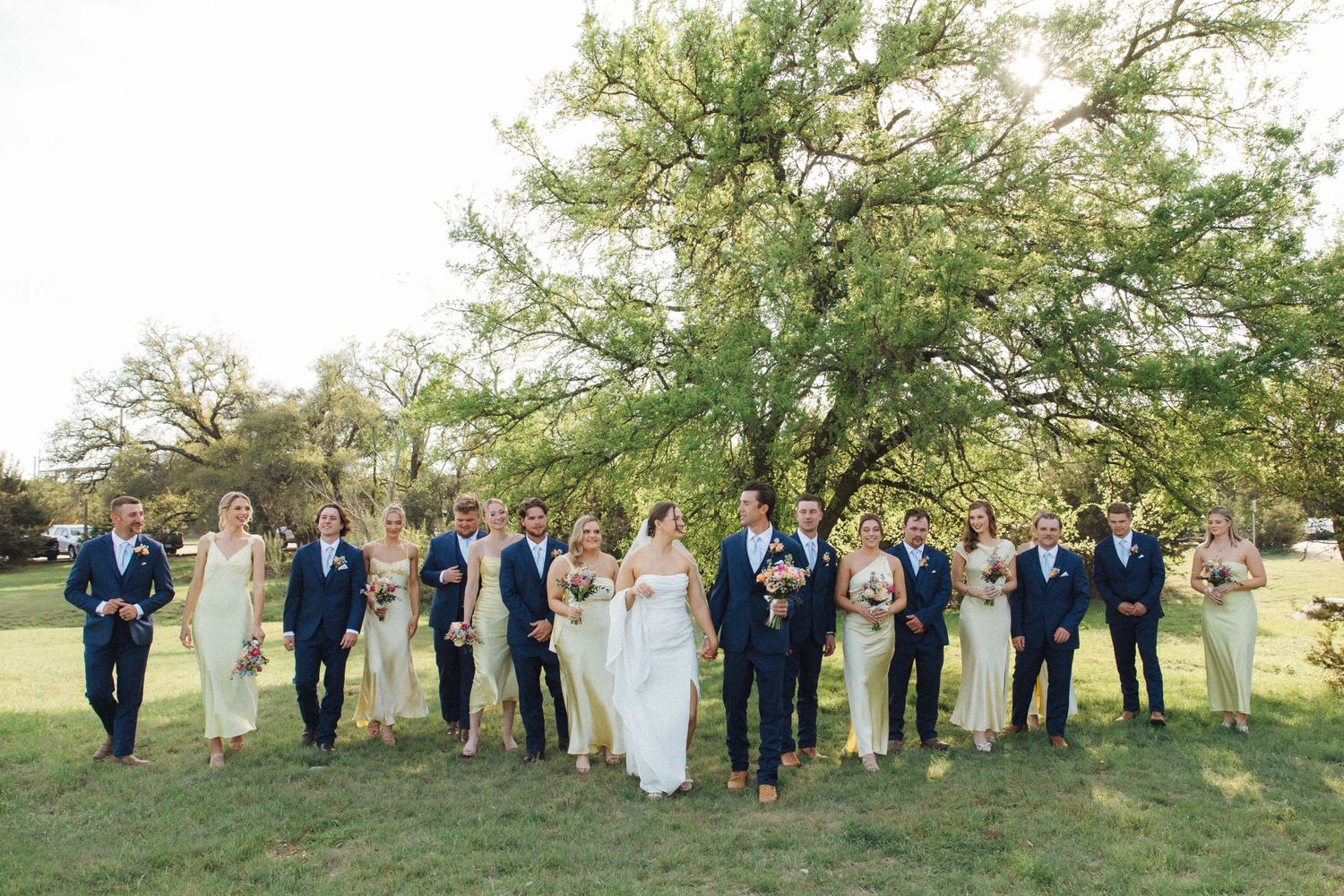 A wedding party in navy suits and formal attire stands together on a grassy hillside for group photos.