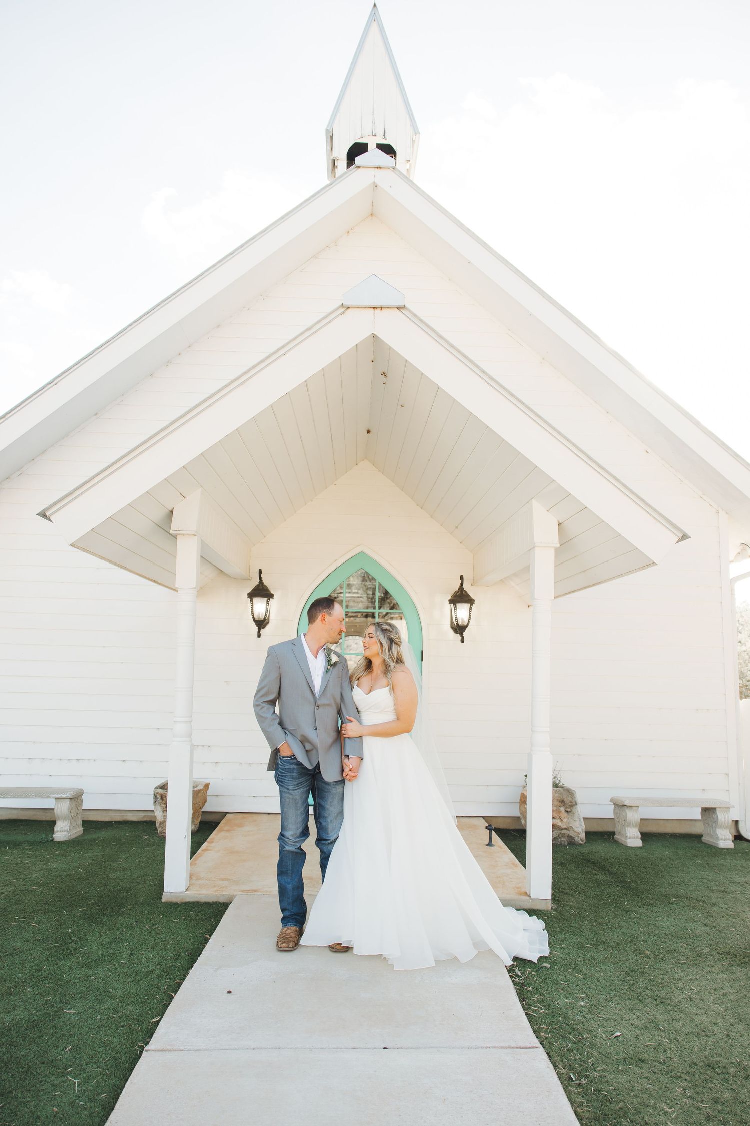 White wooden chapel with Gothic arch doorway and lanterns on a small hill with green grass and concrete pathway.