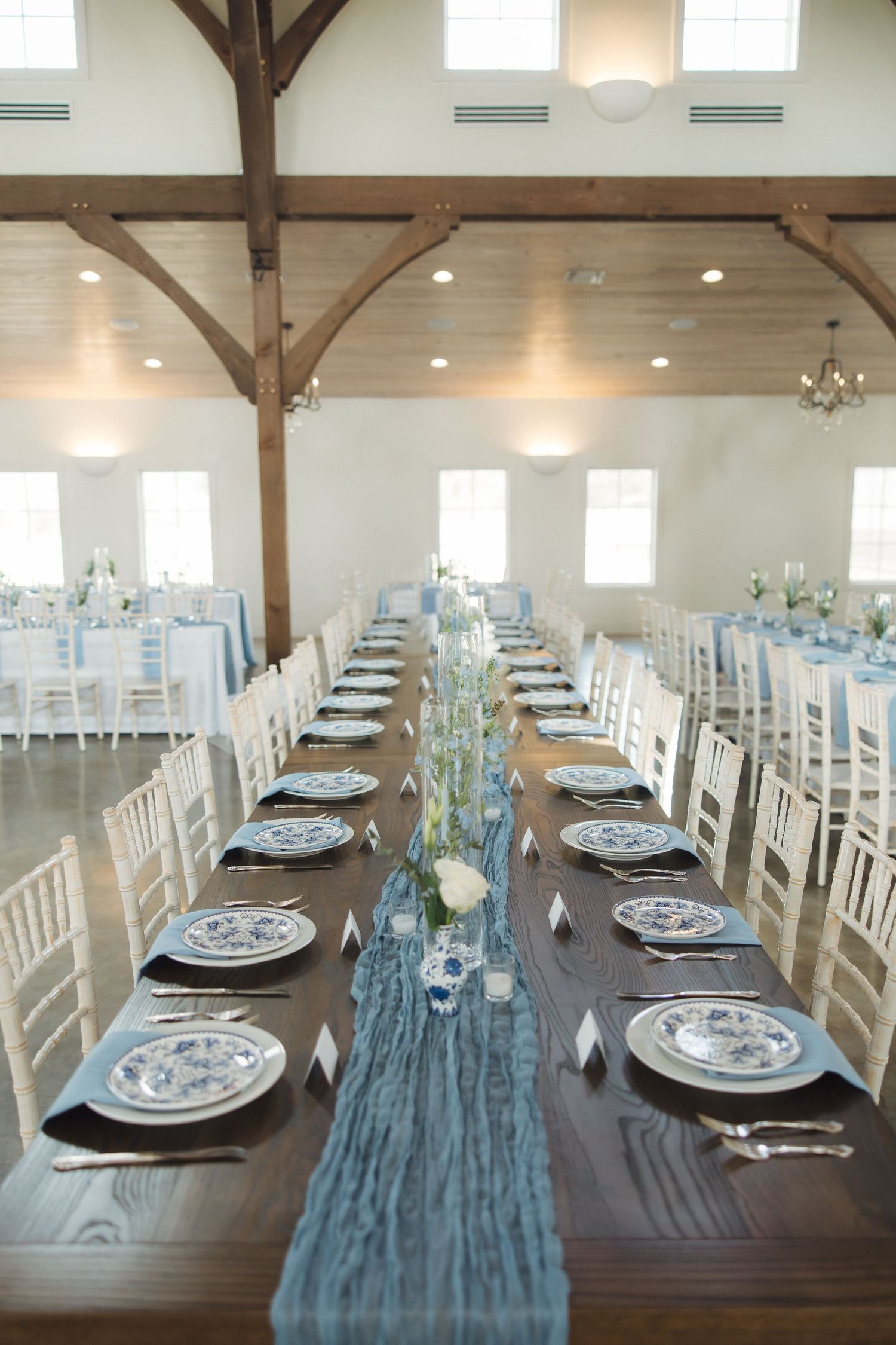 Long rustic wooden tables set for wedding reception with blue runner and white china place settings in modern barn venue.