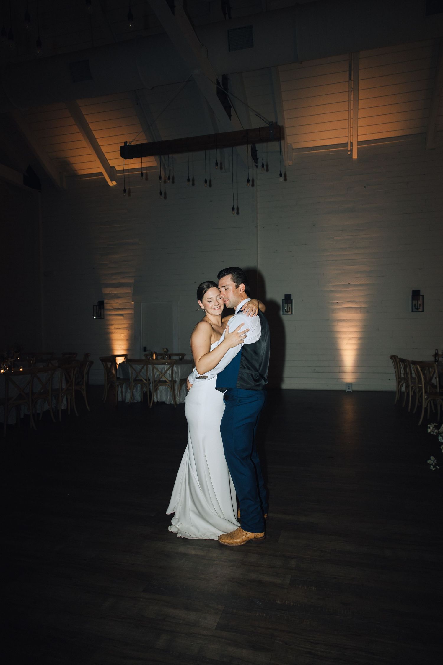 A couple shares their first dance at a wedding reception in a dimly lit venue with warm uplighting on the walls.