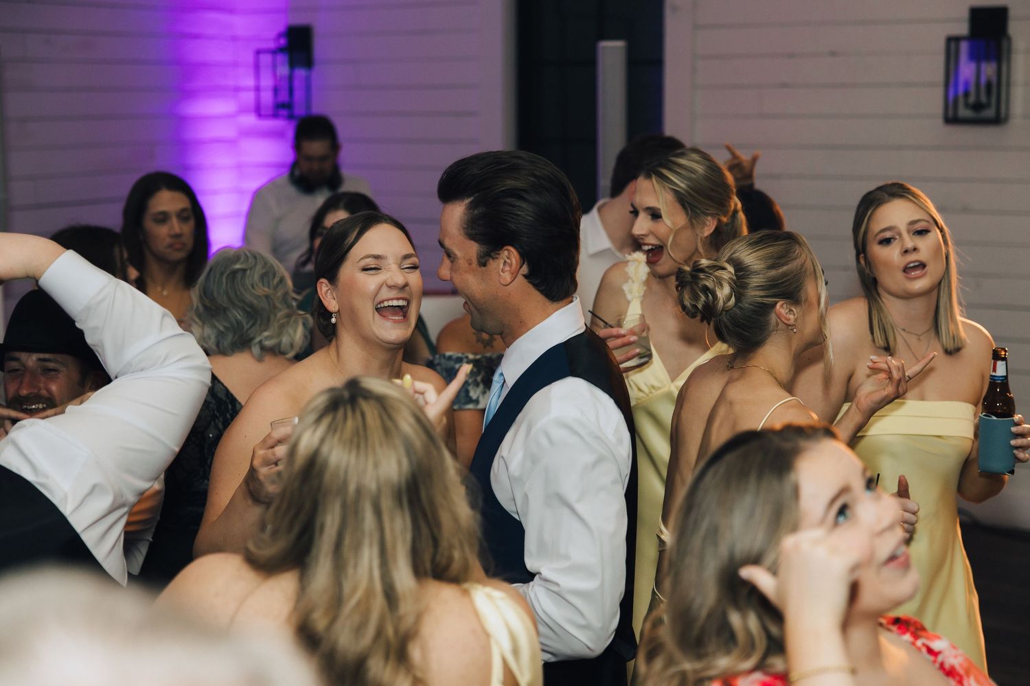 Party guests dancing and celebrating together under purple lighting on a crowded dance floor at an evening event.