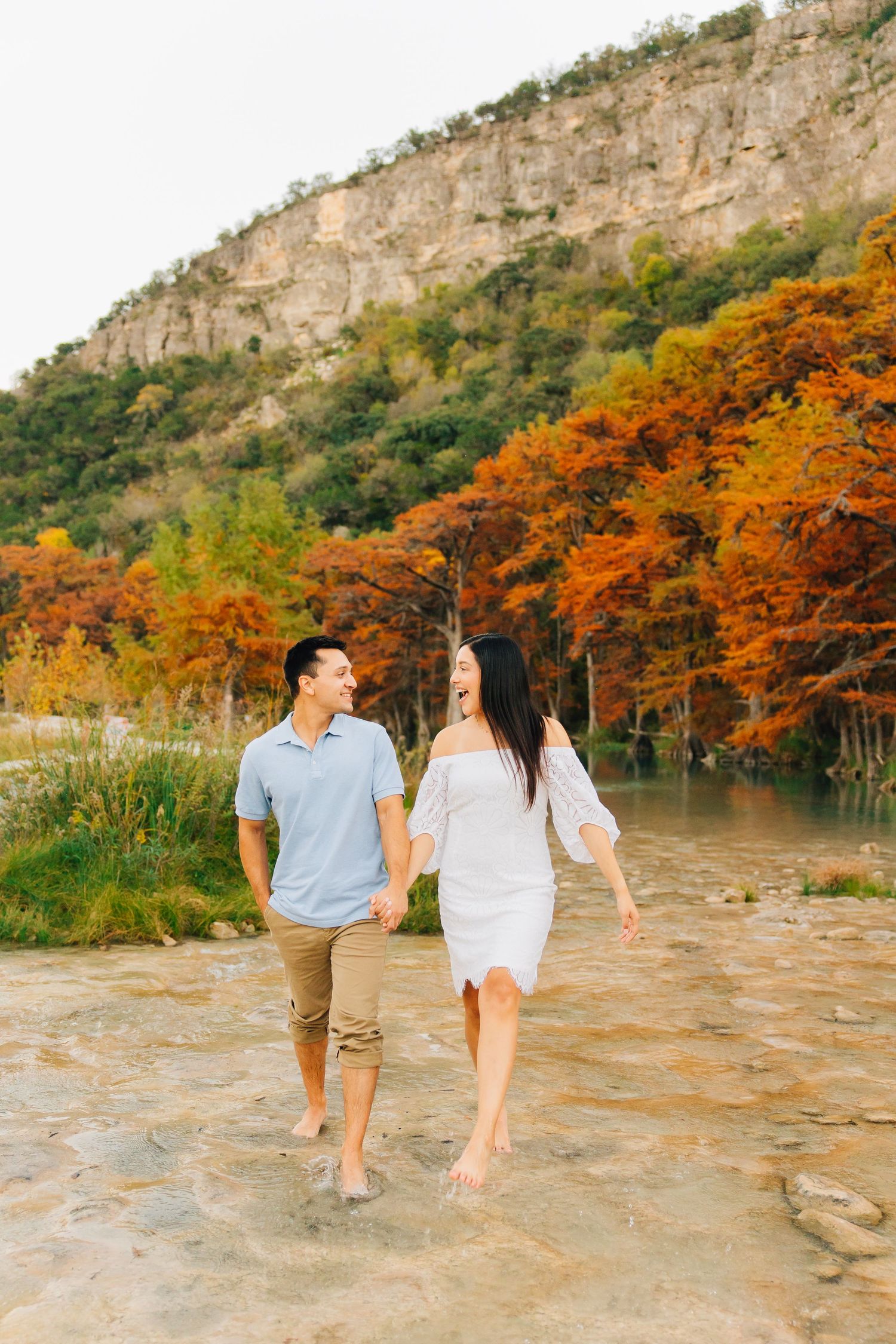 Two people walking barefoot along rocky riverbank with autumn foliage and limestone cliffs in the background.